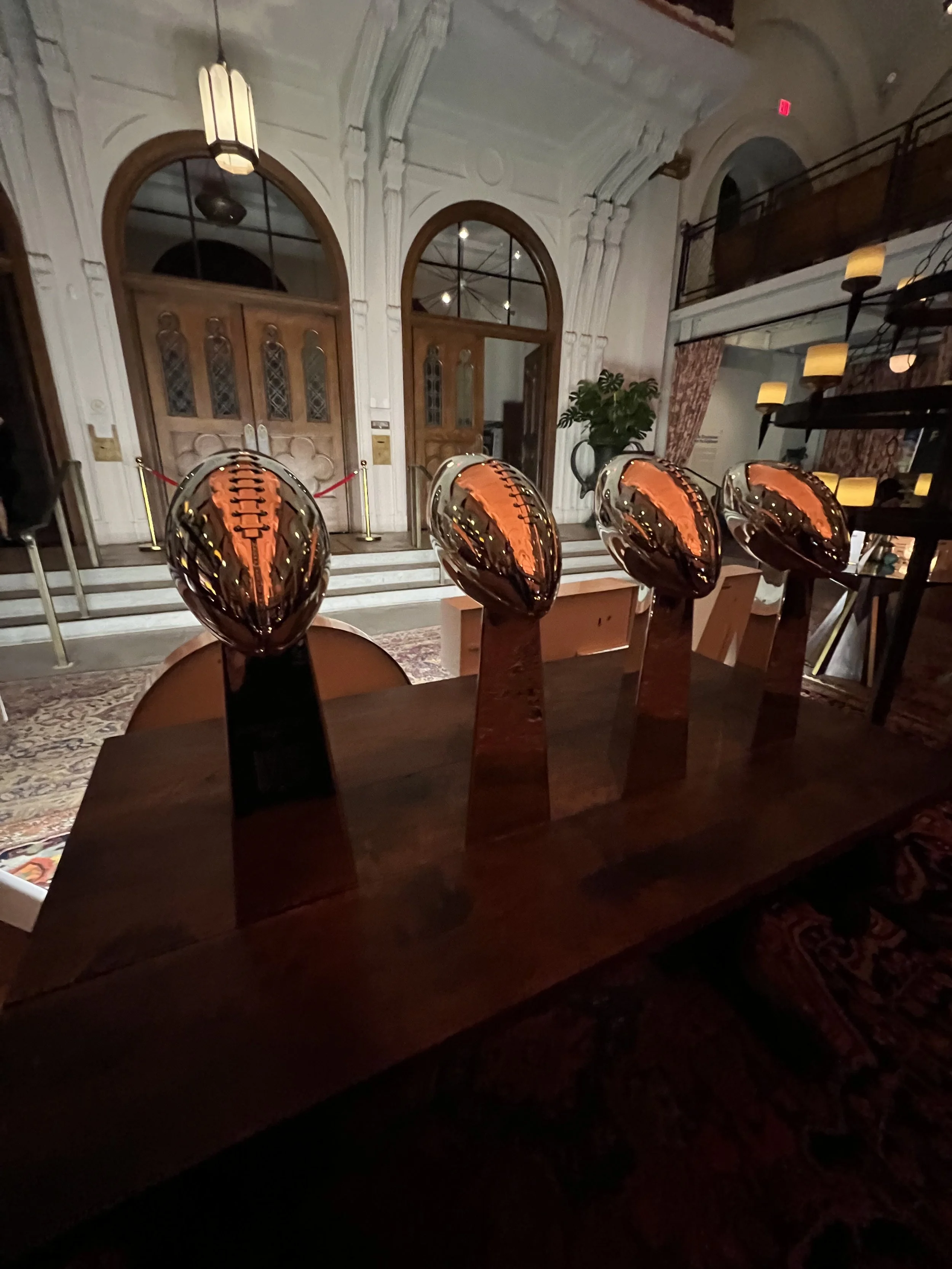 Four shiny football-shaped sculptures on a wooden table in an ornate room with wooden doors, arched windows, and decorative lighting.
