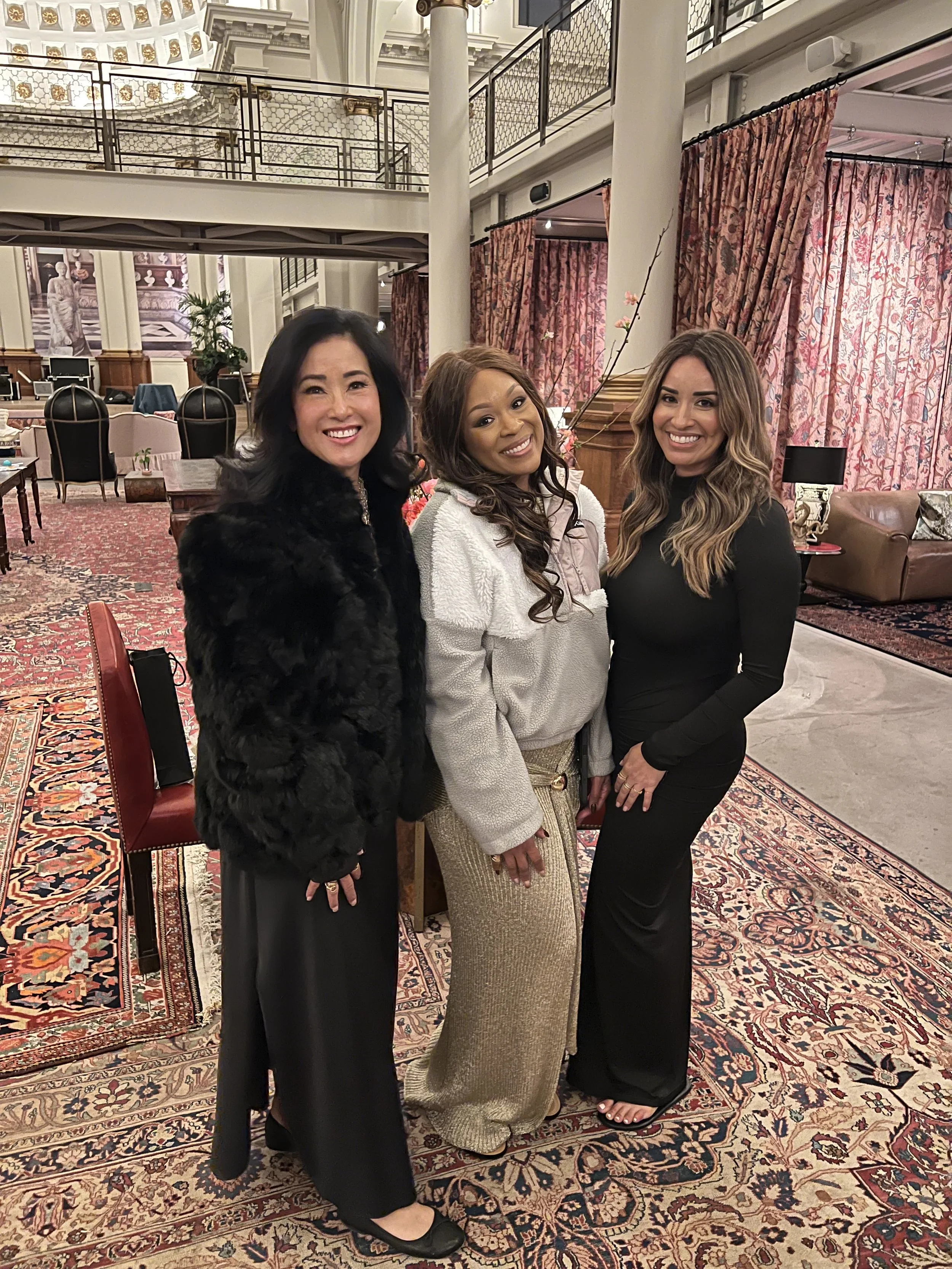 Three women smiling and standing together in an elegant, vintage-style room with ornate carpets, pink curtains, and wooden furniture.