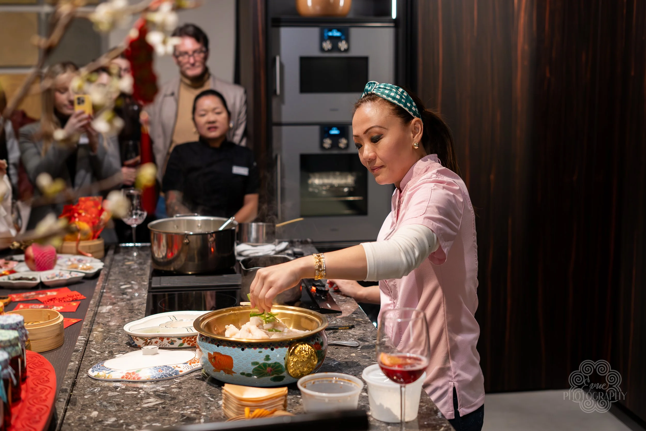 A woman prepares food in a kitchen during a gathering with several people watching her. She is placing ingredients into a decorative bowl, with a wine glass and various dishes on the counter.