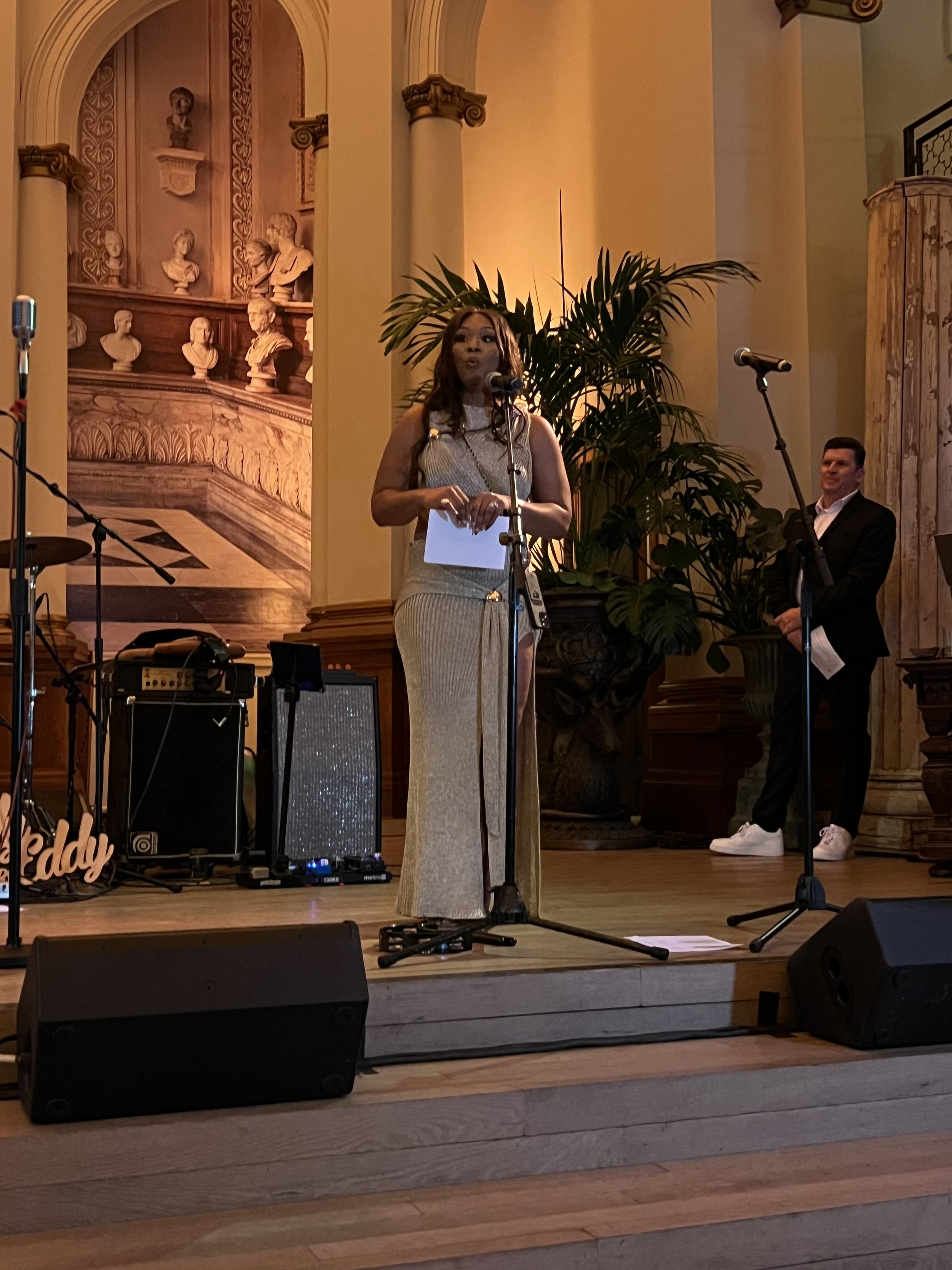 A woman in a gold dress standing at a microphone on a stage, holding papers, with a man in a black suit and white sneakers standing behind her. The stage has musical equipment and a large potted plant, with decorative sculptures and columns in the ba
