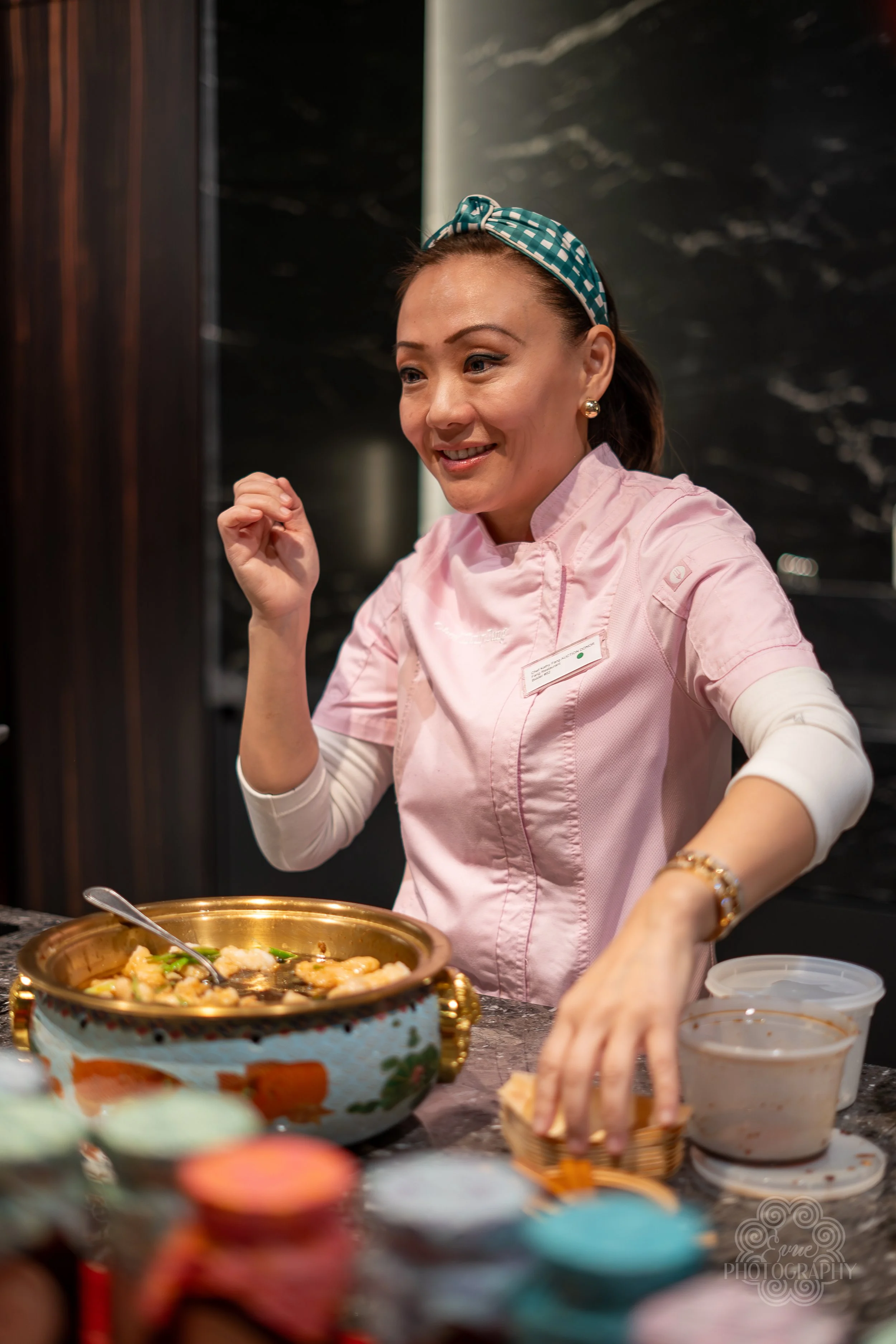 A woman in a pink chef's jacket with a headband and earrings, smiling while preparing food in a kitchen, with a large steaming bowl and various containers on the counter.