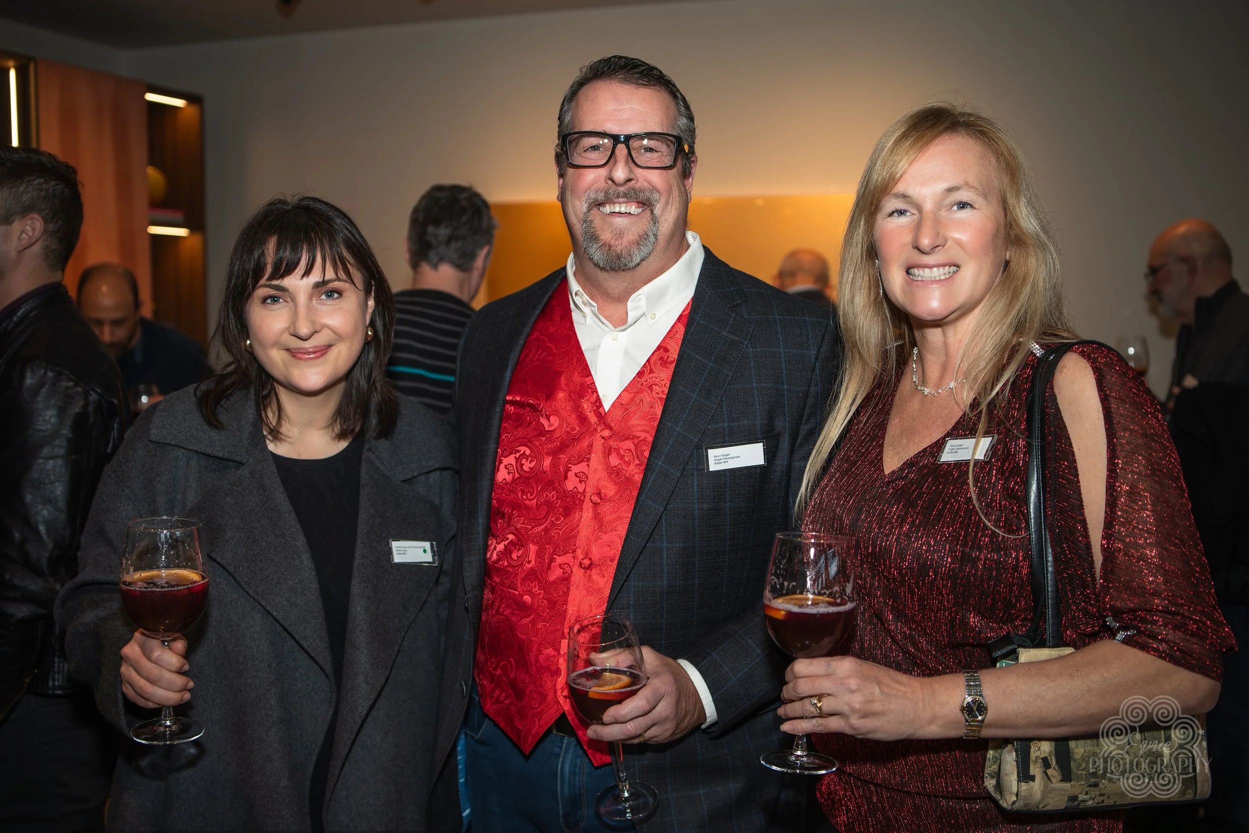 Three people at a social event holding glasses of red wine, smiling at the camera, with a blurred crowd in the background.