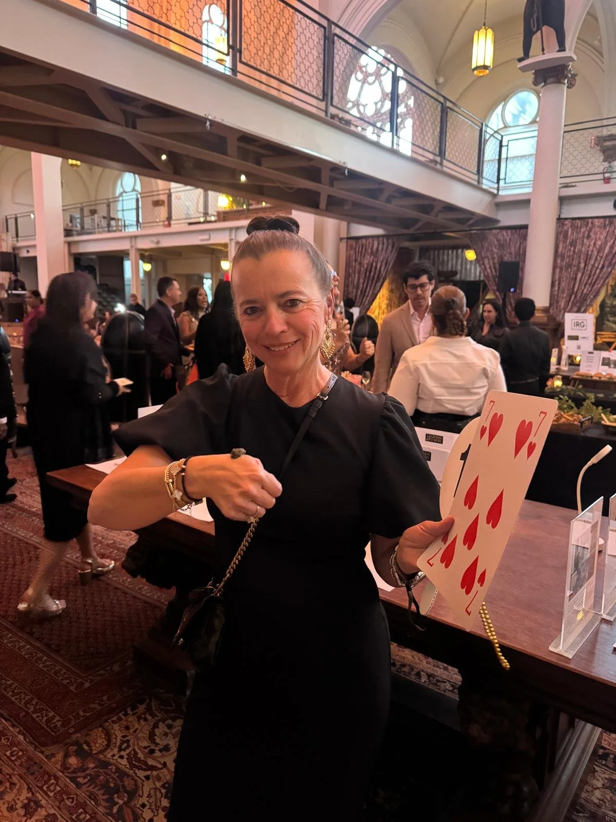 A woman in a black dress holding a large playing card (7 of hearts) at an indoor event with many people in the background.