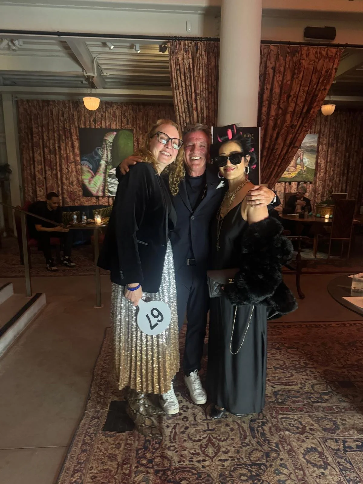 Three people are standing together in a warmly lit indoor setting, smiling and posing for the photo. The person on the left is wearing glasses, a black jacket, and a shiny, metallic pleated skirt. The person in the middle is dressed in a dark suit. T