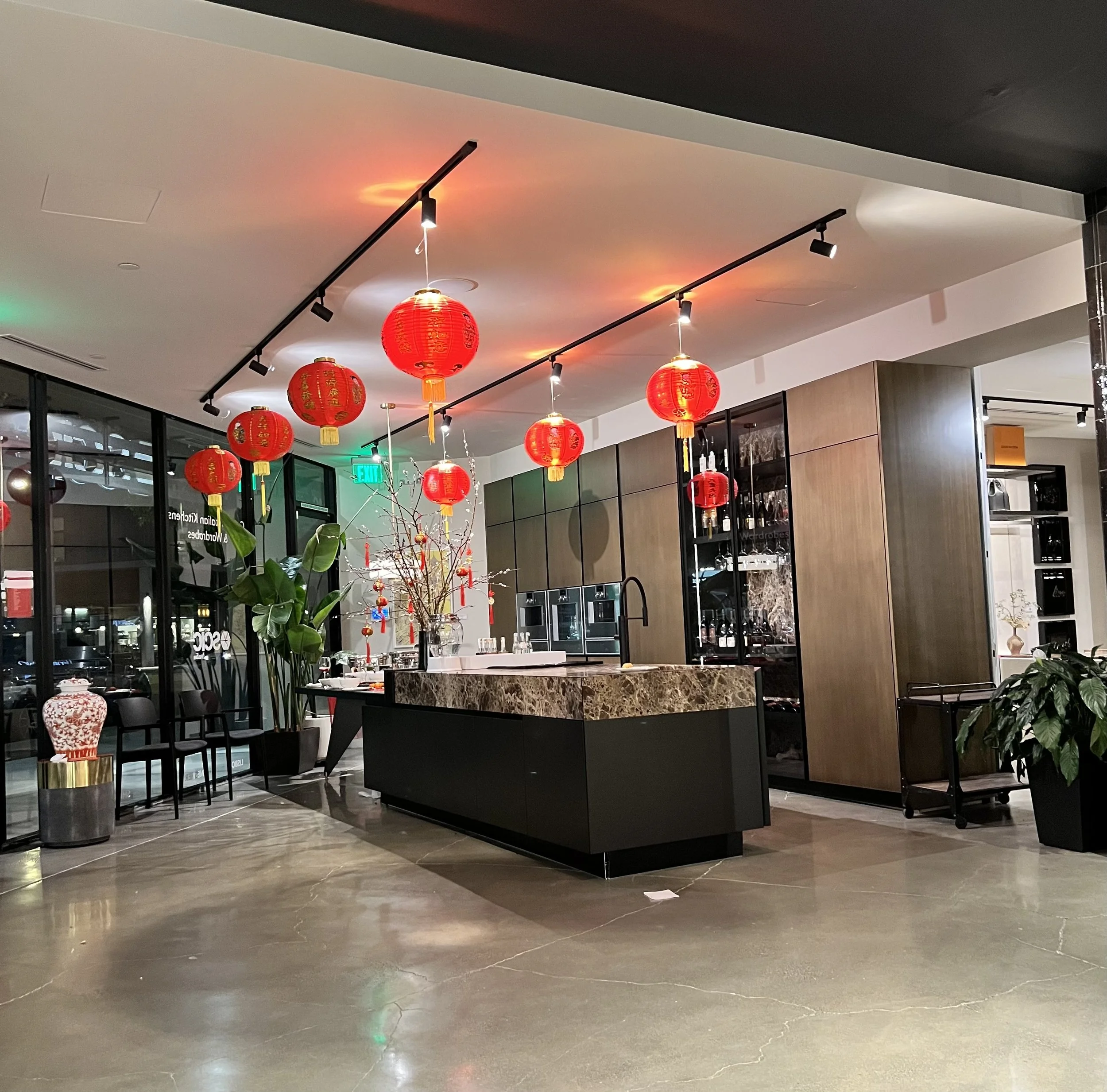 Modern restaurant interior with a black island counter topped with marble, decorated with a branch and Chinese lanterns hanging from the ceiling, large plants, and a seating area near windows.