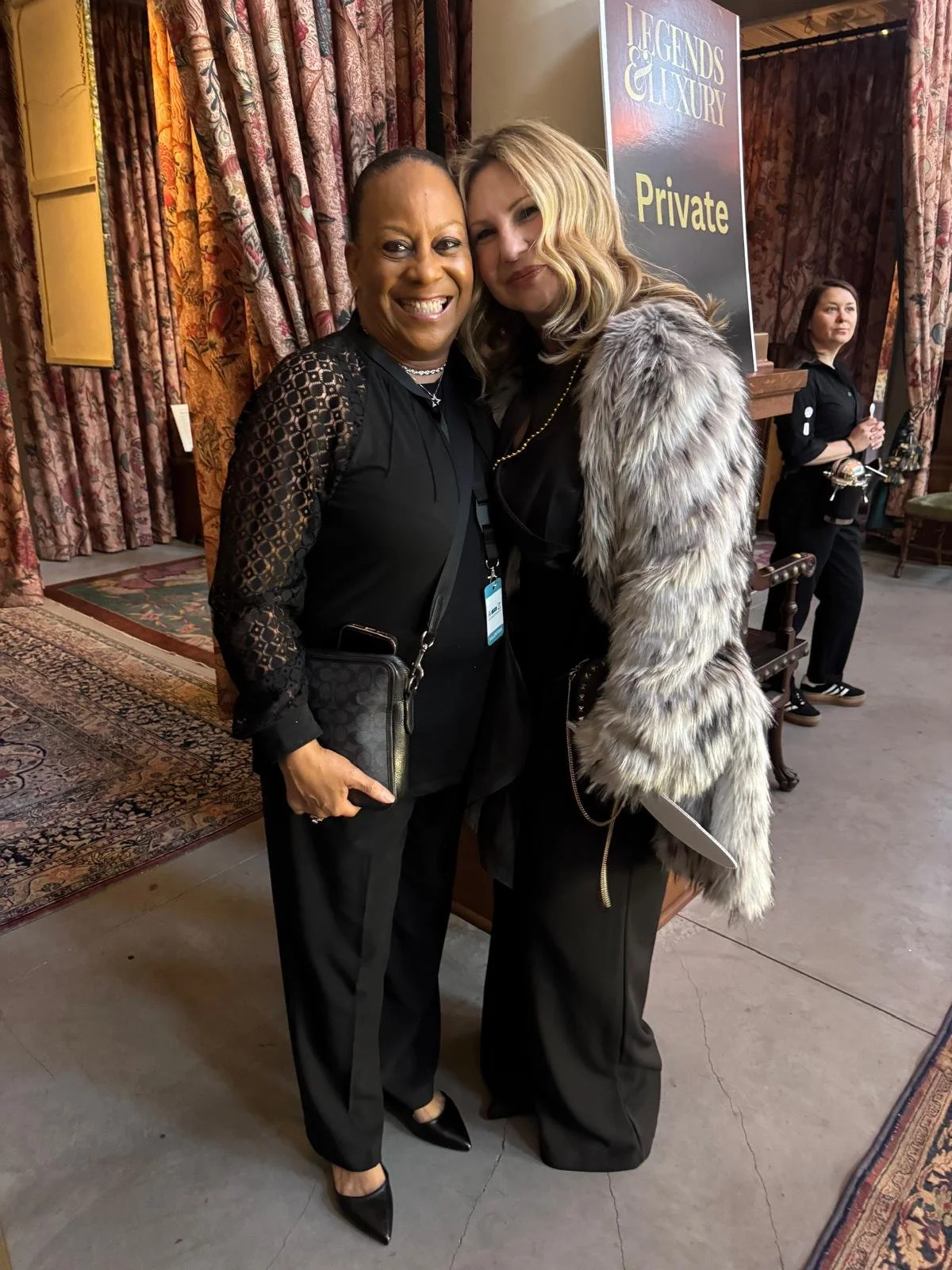 Two women smiling and posing together at an event, one wearing black with lace sleeves and the other dressed in a fur coat, in front of a sign that reads 'Legends & Luxury' and 'Private'.