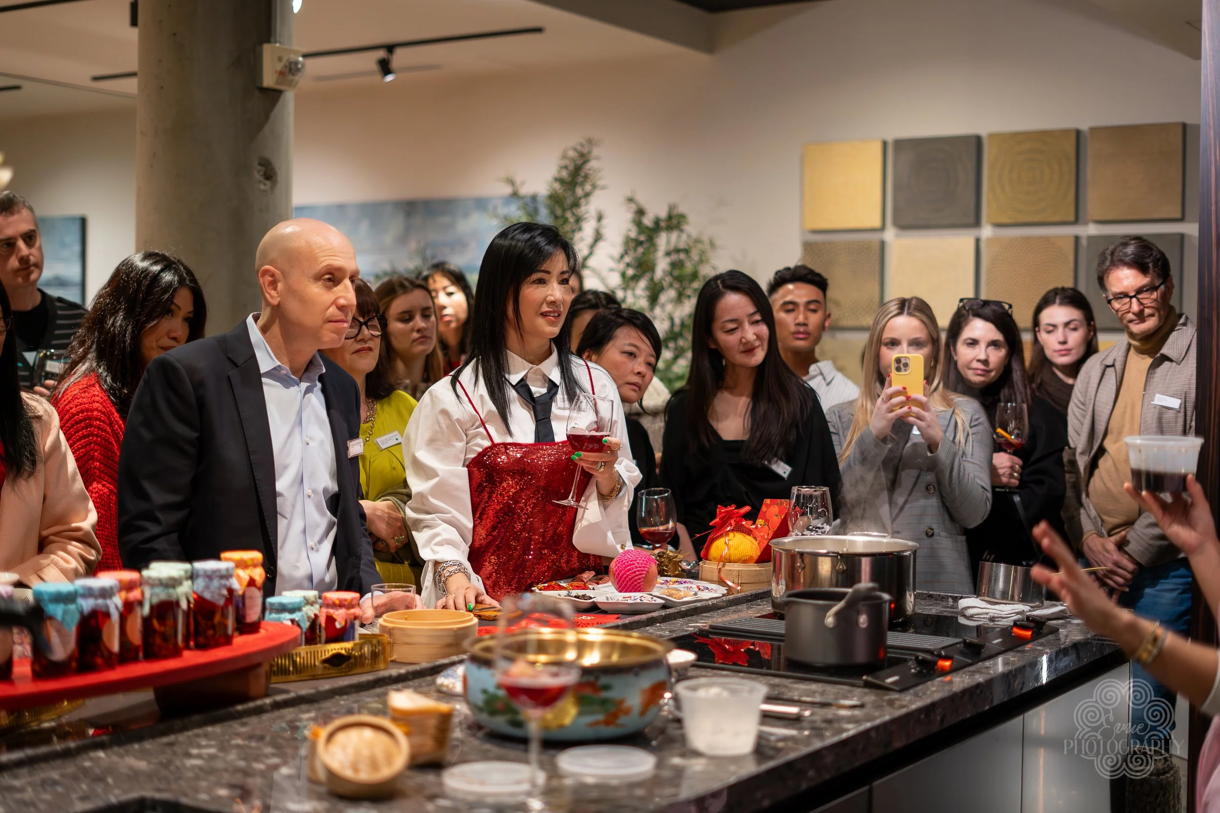 Group of people gathered around a cooking demonstration at a party or event, with a woman in a red apron leading the activity, on a kitchen island with food and drinks, taking photos and watching intently.