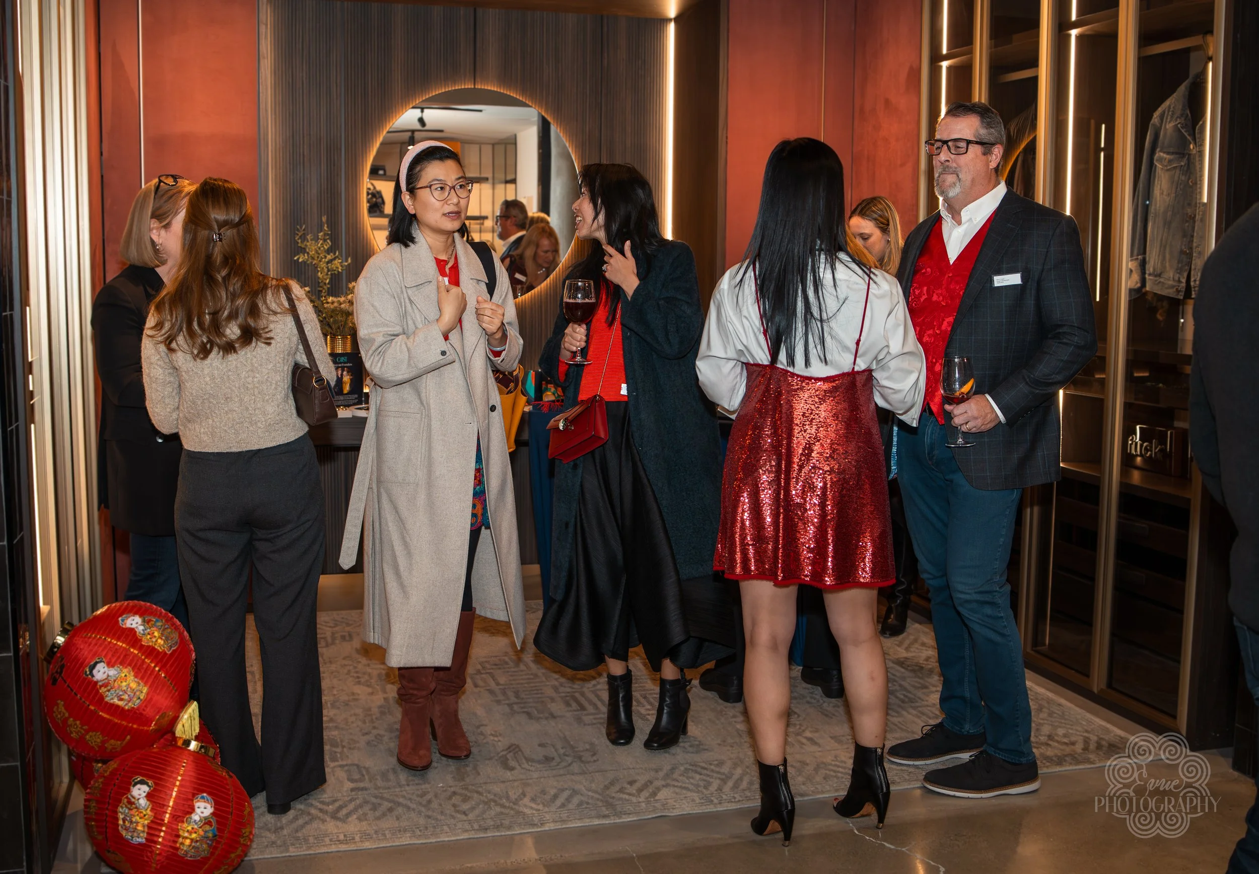 Group of people chatting at a social gathering in a trendy indoor venue, with warm lighting, modern decor, and a mix of casual and semi-formal attire, including women in dresses and men in blazers, some holding glasses of wine.