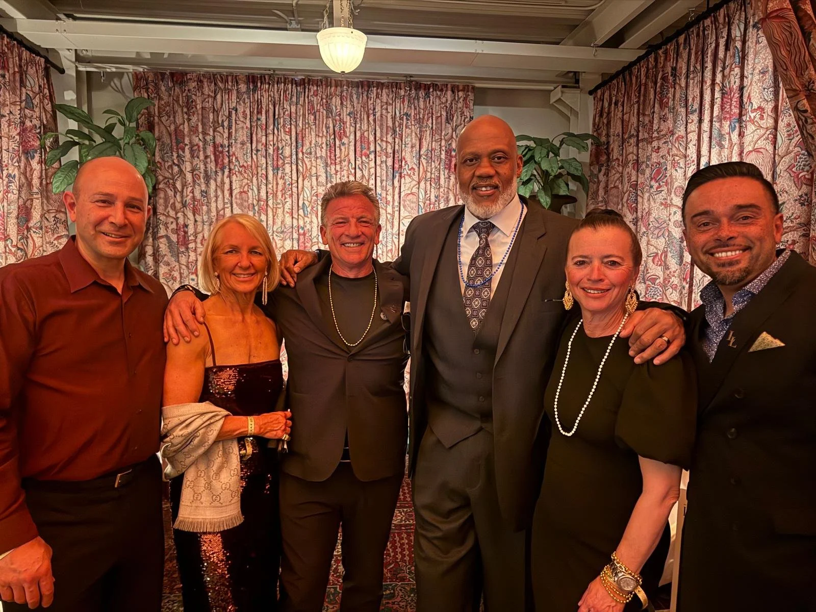 Group of six adults smiling, standing close together in a room with floral curtains and plants.