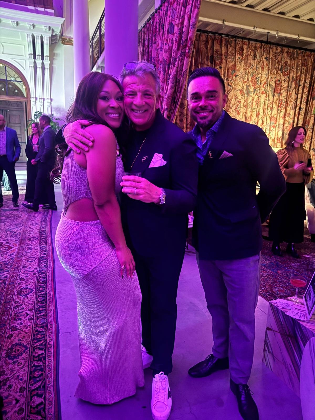 Three people smiling and hugging at an indoor event, with gold curtains and decorative architecture in the background. The woman on the left is wearing a sleeveless, shimmering dress; the man in the middle is holding a drink and wearing a dark blazer