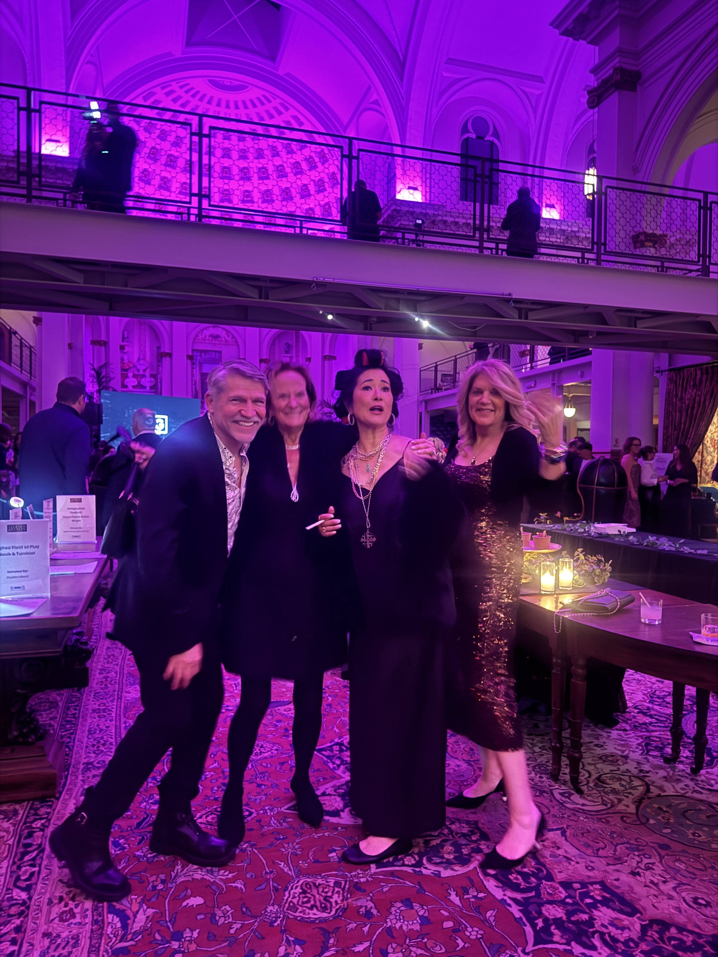Four people standing together at an elegant indoor event with purple lighting, smiling, in a grand hall with ornate decor, tables, and an upstairs balcony.