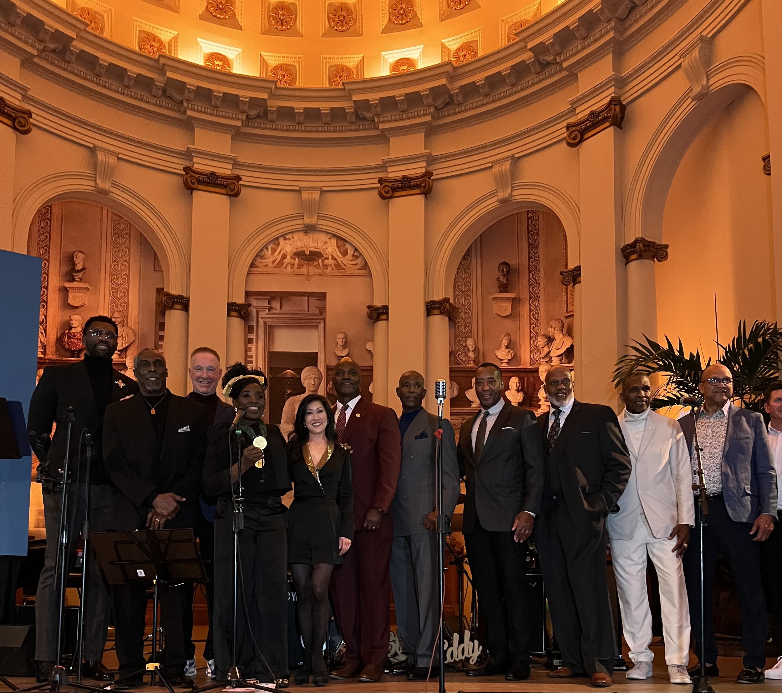 A group of people standing on stage with microphones in front of a classical style, ornate interior with bust sculptures and decorative columns, under warm yellow lighting.