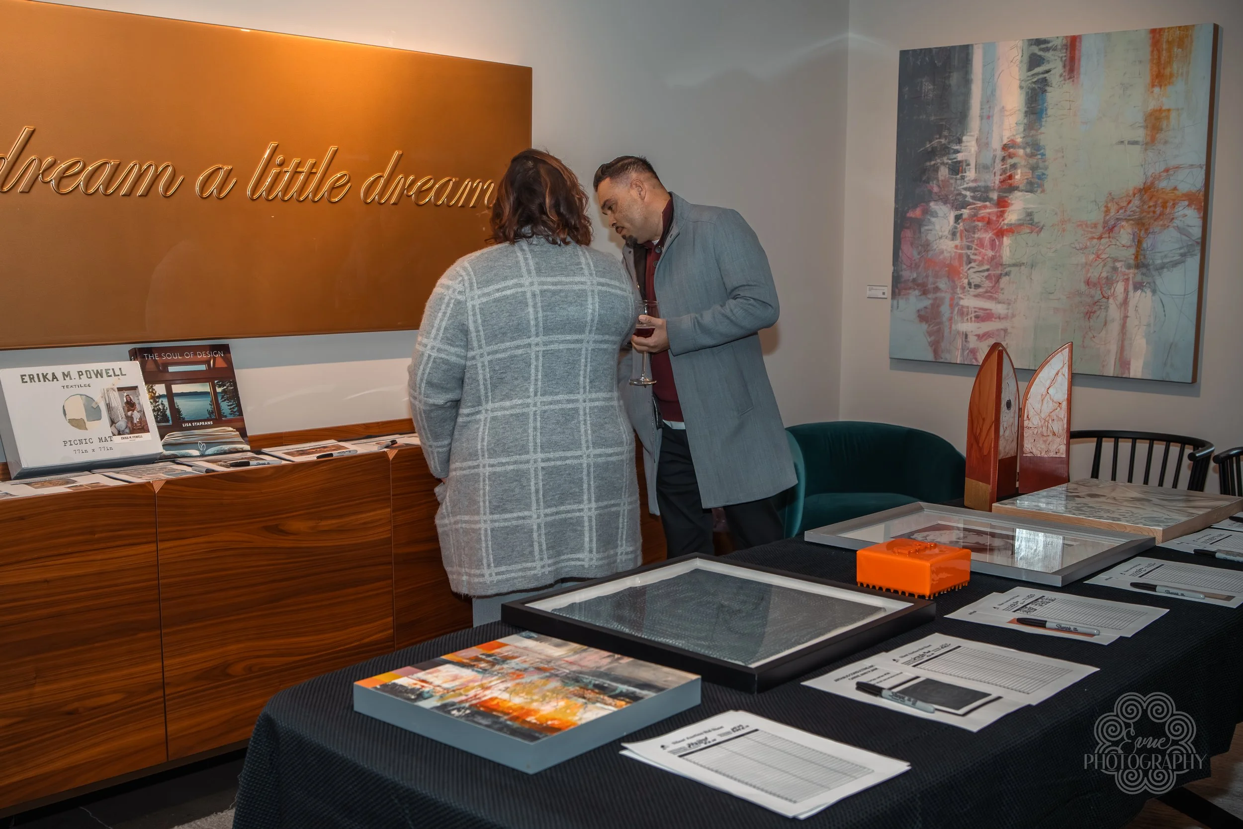 Two people talking at an art gallery, with paintings on the wall and tables with brochures and artwork.