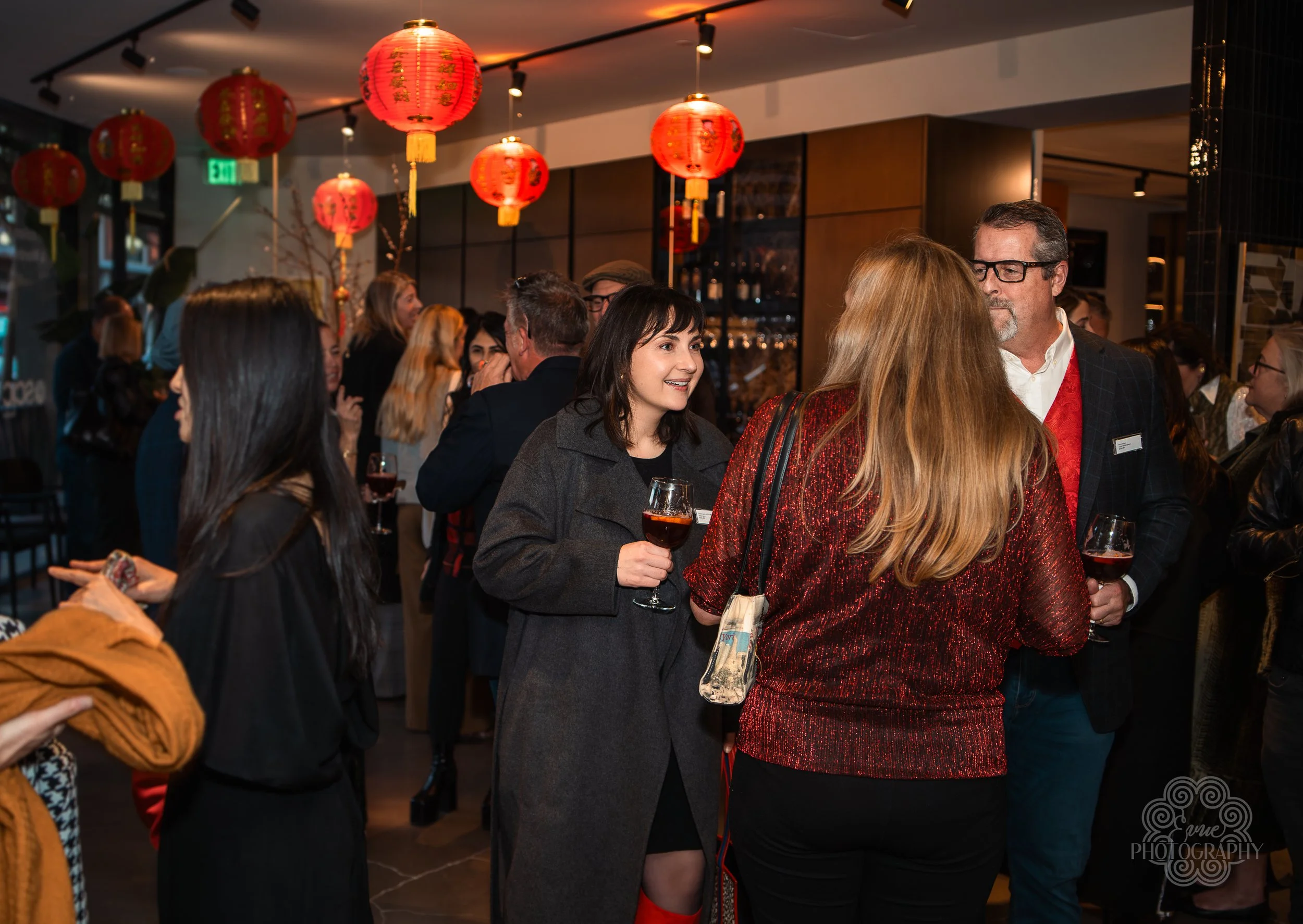People socializing at a festive event decorated with red lanterns, some holding glasses of red wine, in a warmly lit indoor space.