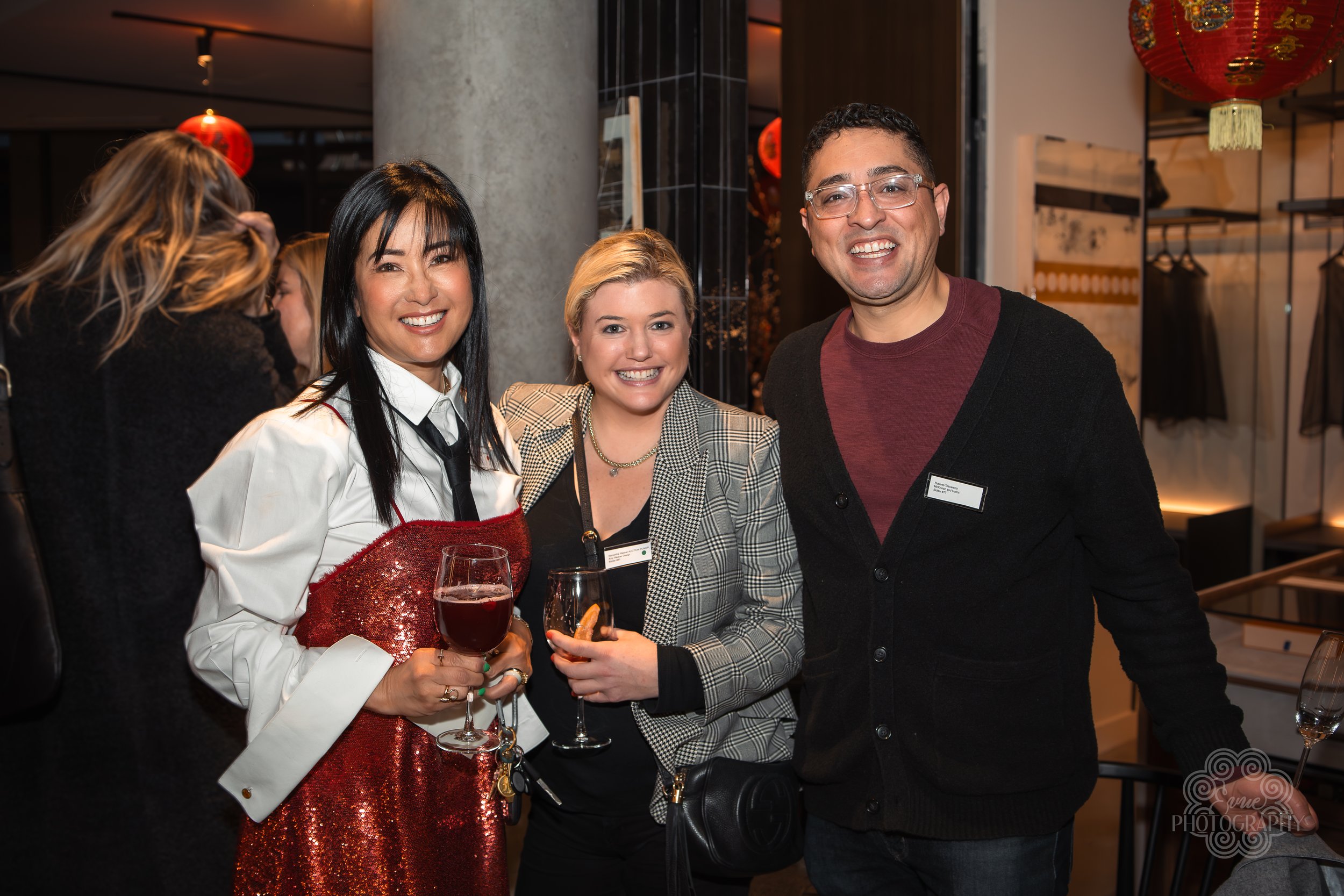 Three people smiling and holding drinks at a social event, with Asian woman in a shiny red dress and black tie, blonde woman in a checkered blazer, and a man in a maroon shirt with glasses.