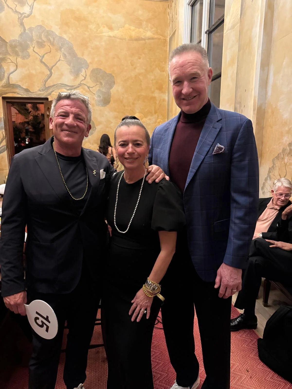 Three people smiling for the camera at an indoor event. The man on the left has gray hair, wearing a black suit with a beaded necklace. The woman in the middle has dark hair pulled back, wearing a black dress with pearl necklace and bracelets. The ma