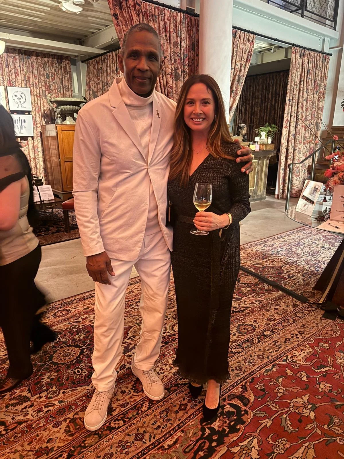 A man in a white suit standing next to a woman in a black dress, both smiling and holding wine glasses at an indoor event with ornate curtains and patterned rugs.
