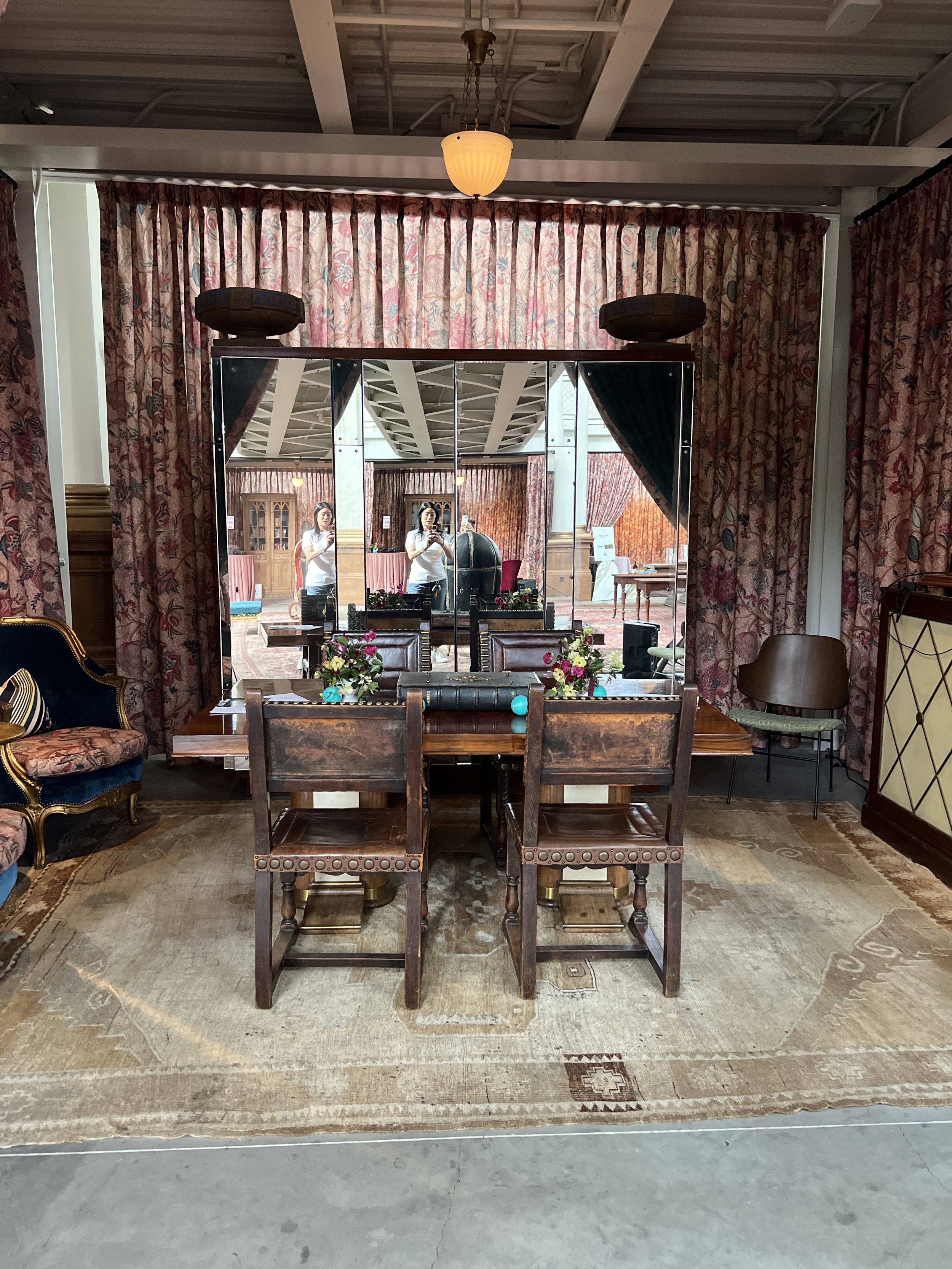 A room with a wooden table and chairs, a large mirror reflecting two women taking a photo, floral curtains, floral arrangements, and vintage furniture.