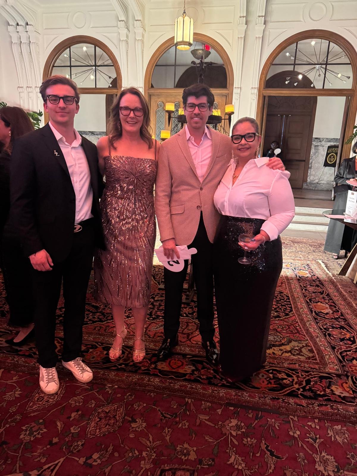 A group of four people dressed in formal attire standing together in an elegant room with ornate white walls and large arched windows, posing for a photo at a social event or gala.