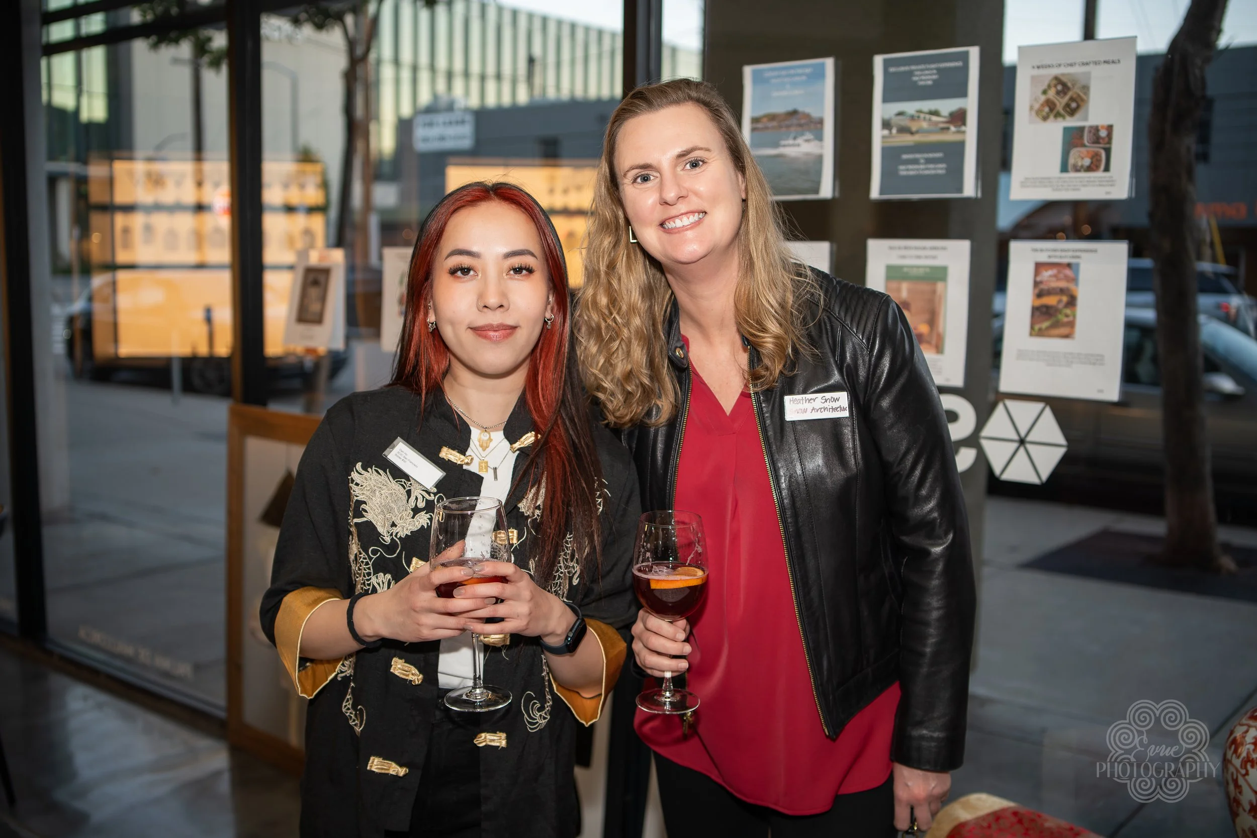 Two women standing inside a modern gallery or event space, holding wine glasses and smiling at the camera. Behind them are display panels with posters, and through large glass windows, outdoor scenery and parked cars are visible.