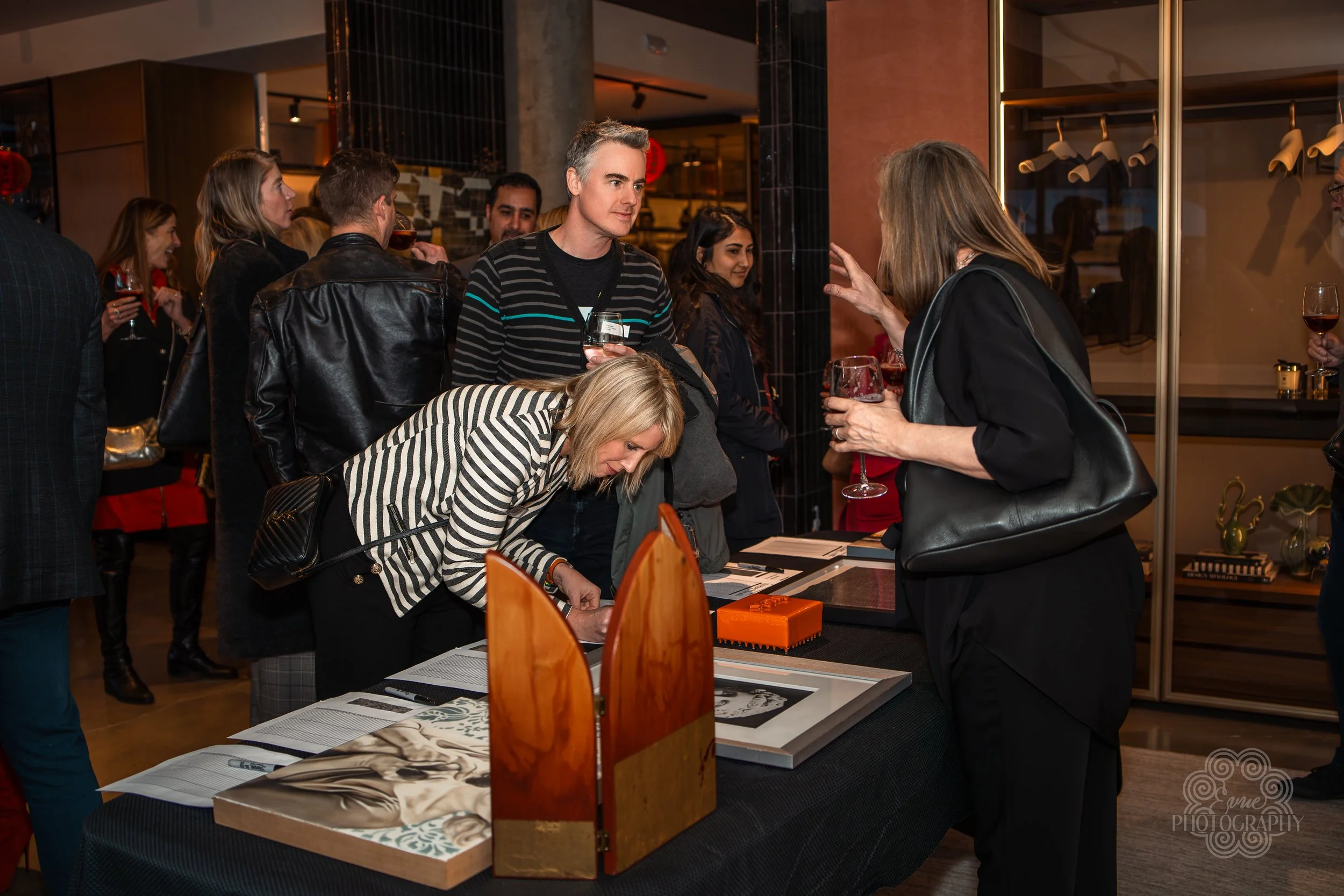 People socializing at an indoor event, displaying artwork on a table, with drinks in hand.