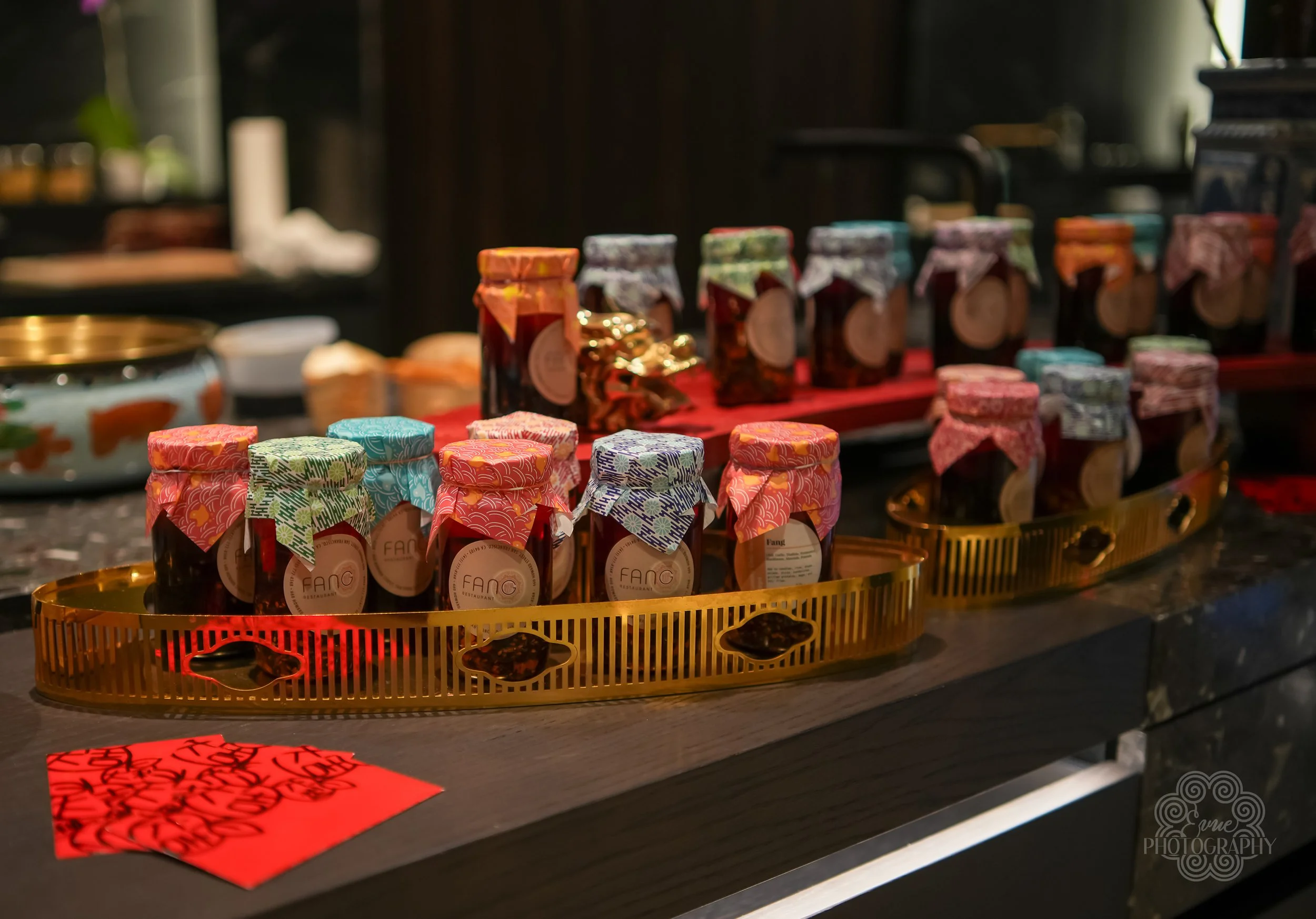 Assorted jars of jam with colorful fabric covers on gold trays, displayed on a dark wooden counter.