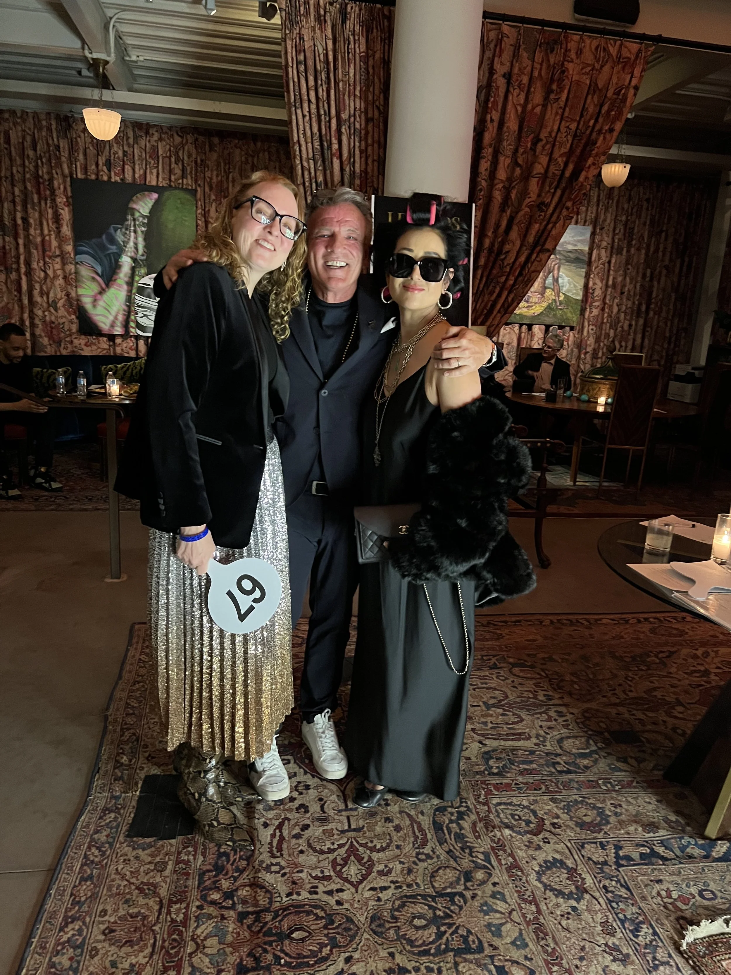 Three people dressed stylishly, posing happily together at an indoor social event. The woman on the left wears glasses, a black blazer, and a glittery skirt, holding a sign with the number 67. The man in the middle has gray hair, wears a dark suit, a