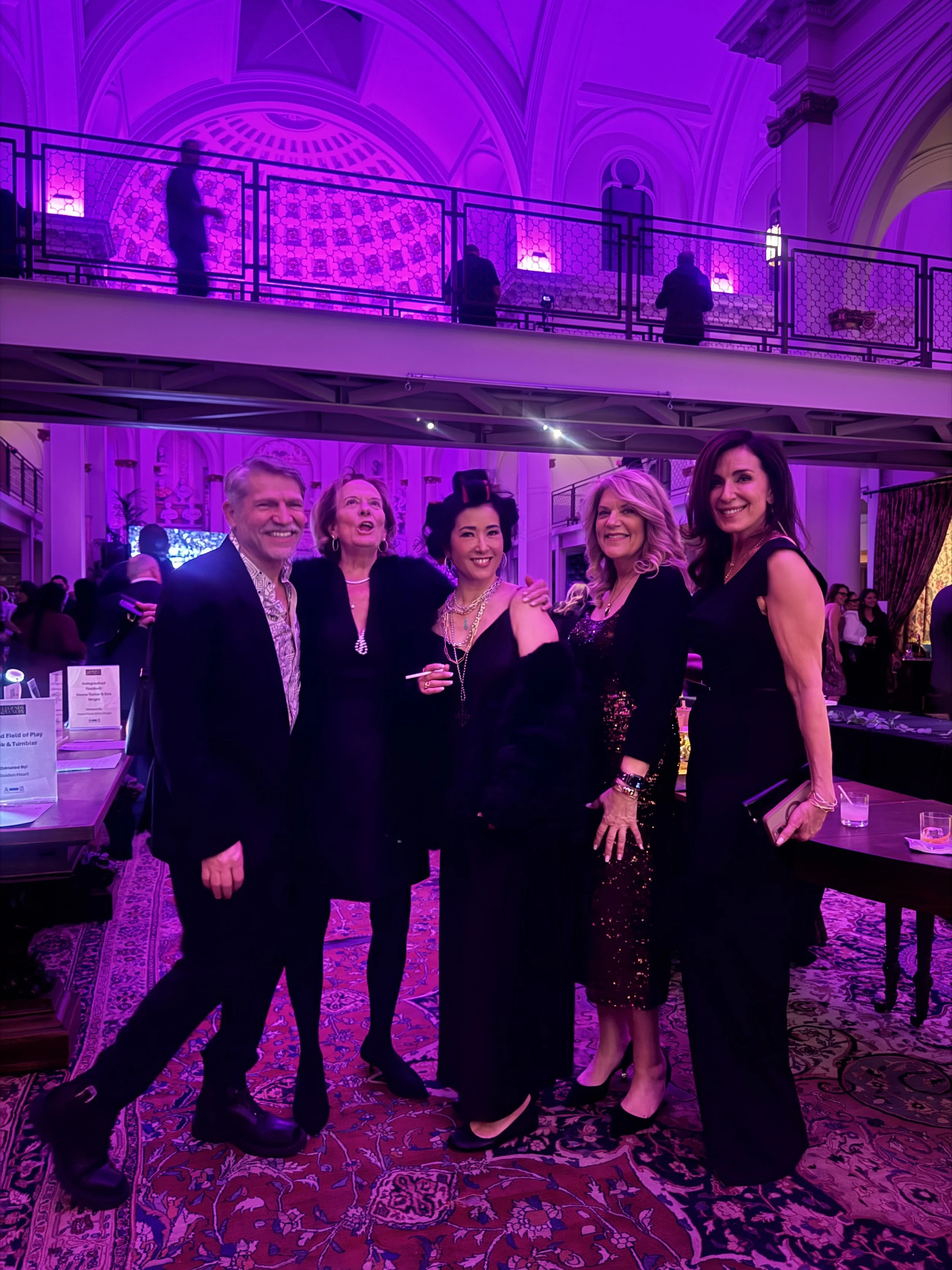 Group of five people dressed in formal attire at an indoor event with purple lighting, standing on a patterned carpet in a large, ornate hall.