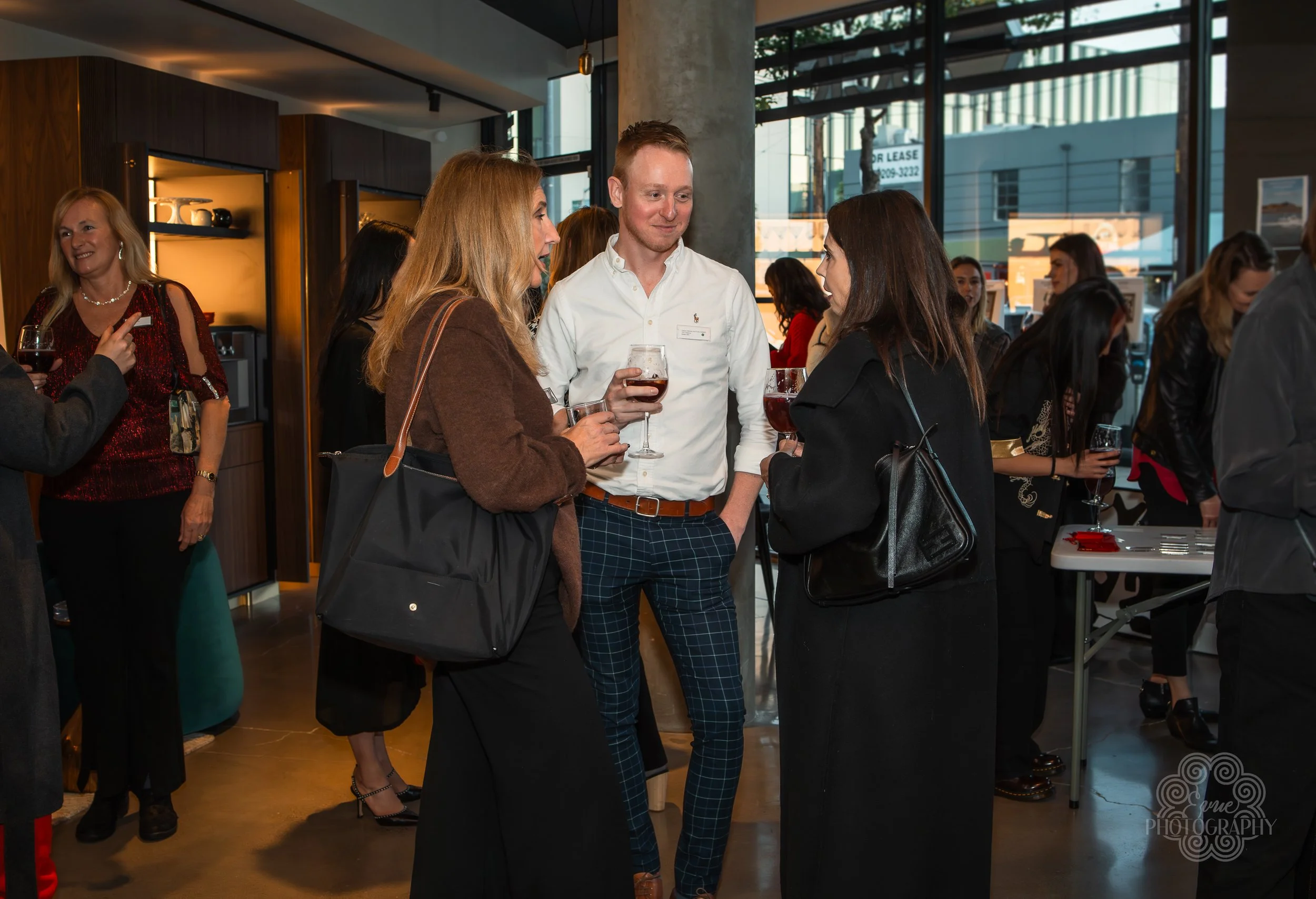 Group of people socializing indoors at a networking event, holding glasses of wine and engaging in conversation.