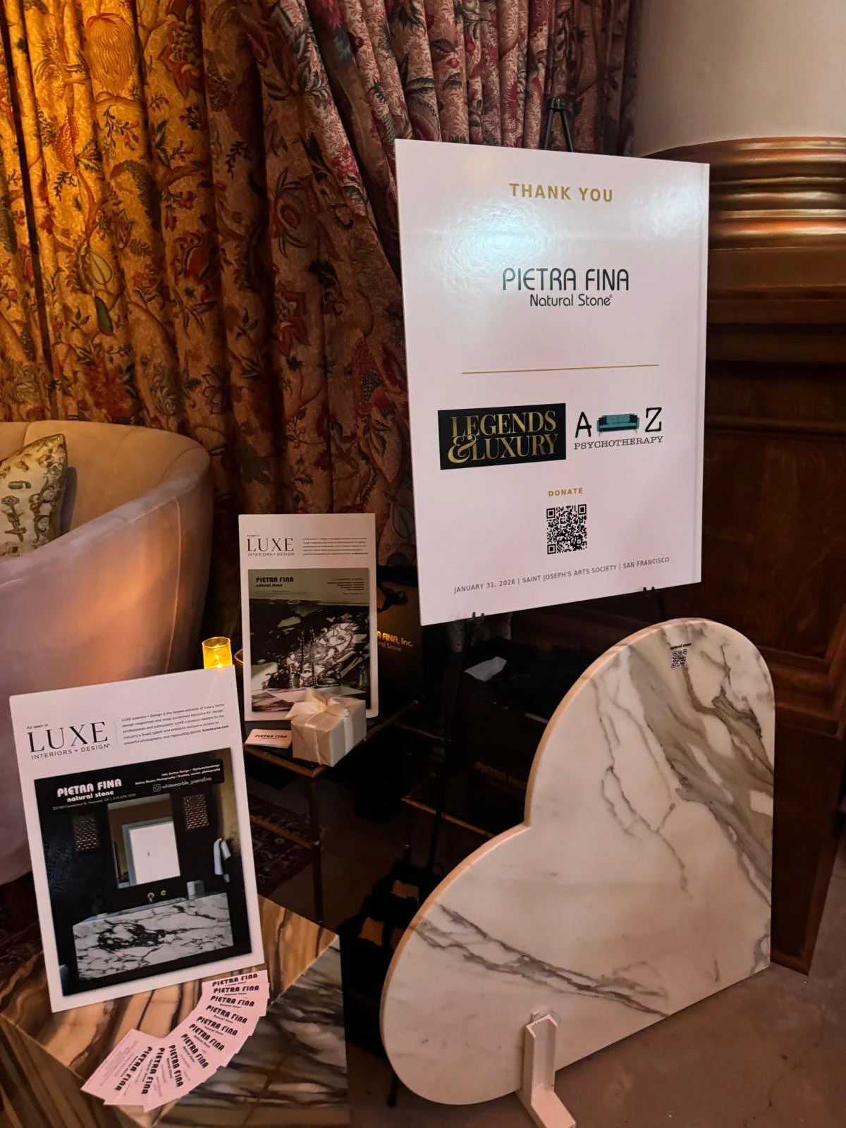 Display of promotional materials for Pietra Fina Natural Stone at an event, featuring a large white poster with gold and black text, smaller flyers, and a marble sample block set against a backdrop of ornate curtains and furniture.