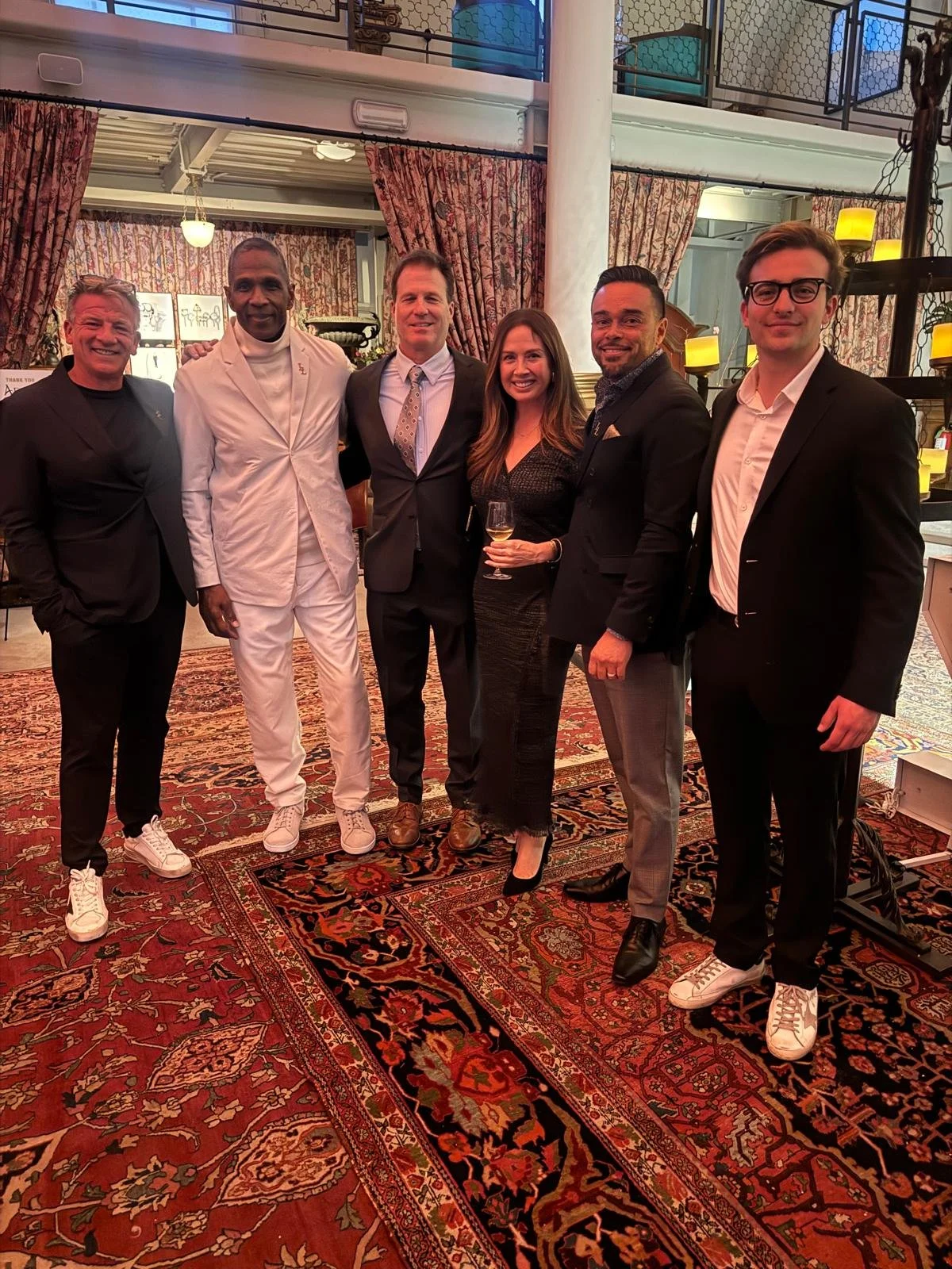 Group of six people dressed in formal and semi-formal attire posing together indoors at an event, with a decorative rug and vintage curtains in the background.