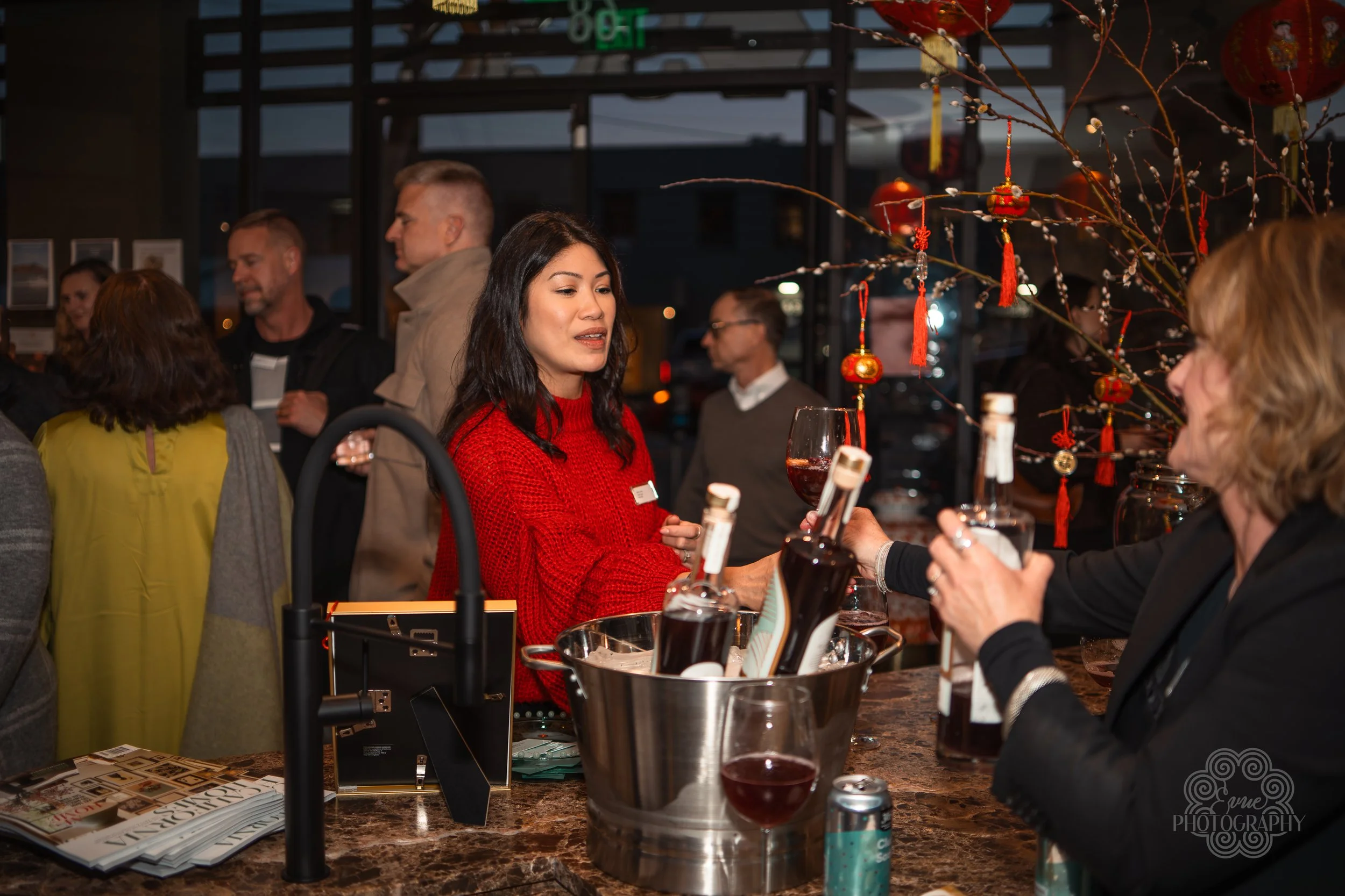 A woman in a red sweater at a bar or reception area, receiving a drink from another woman. The background shows a gathering of people in a decorated interior, possibly for a celebration or event.