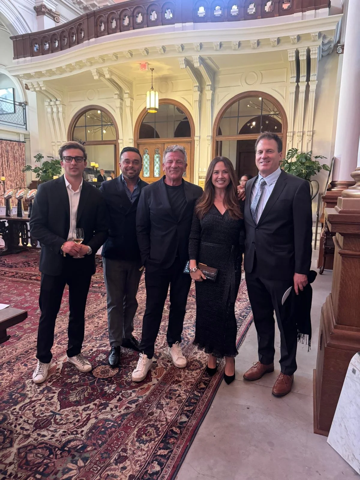 A group of five people dressed in formal attire standing together inside a grand, elegantly decorated building with arched windows, ornate molding, and a vintage carpet.