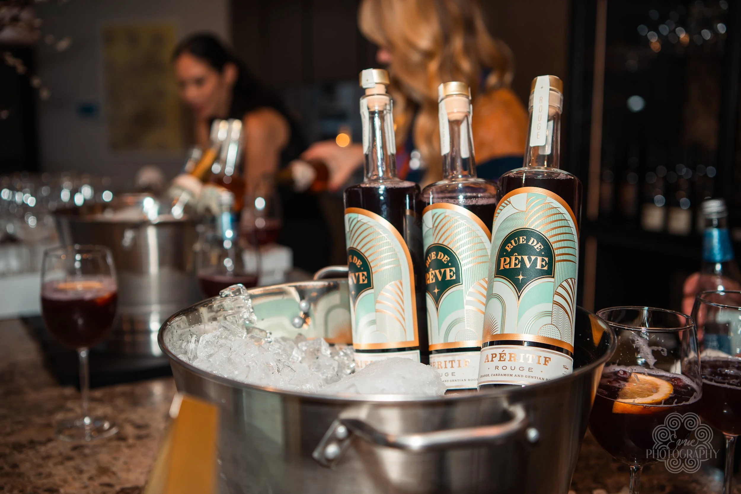 Bottles of Rue de Rêve Aperitif Rouge in a bucket with ice, surrounded by glasses of red wine, with two women in the background at a bar.