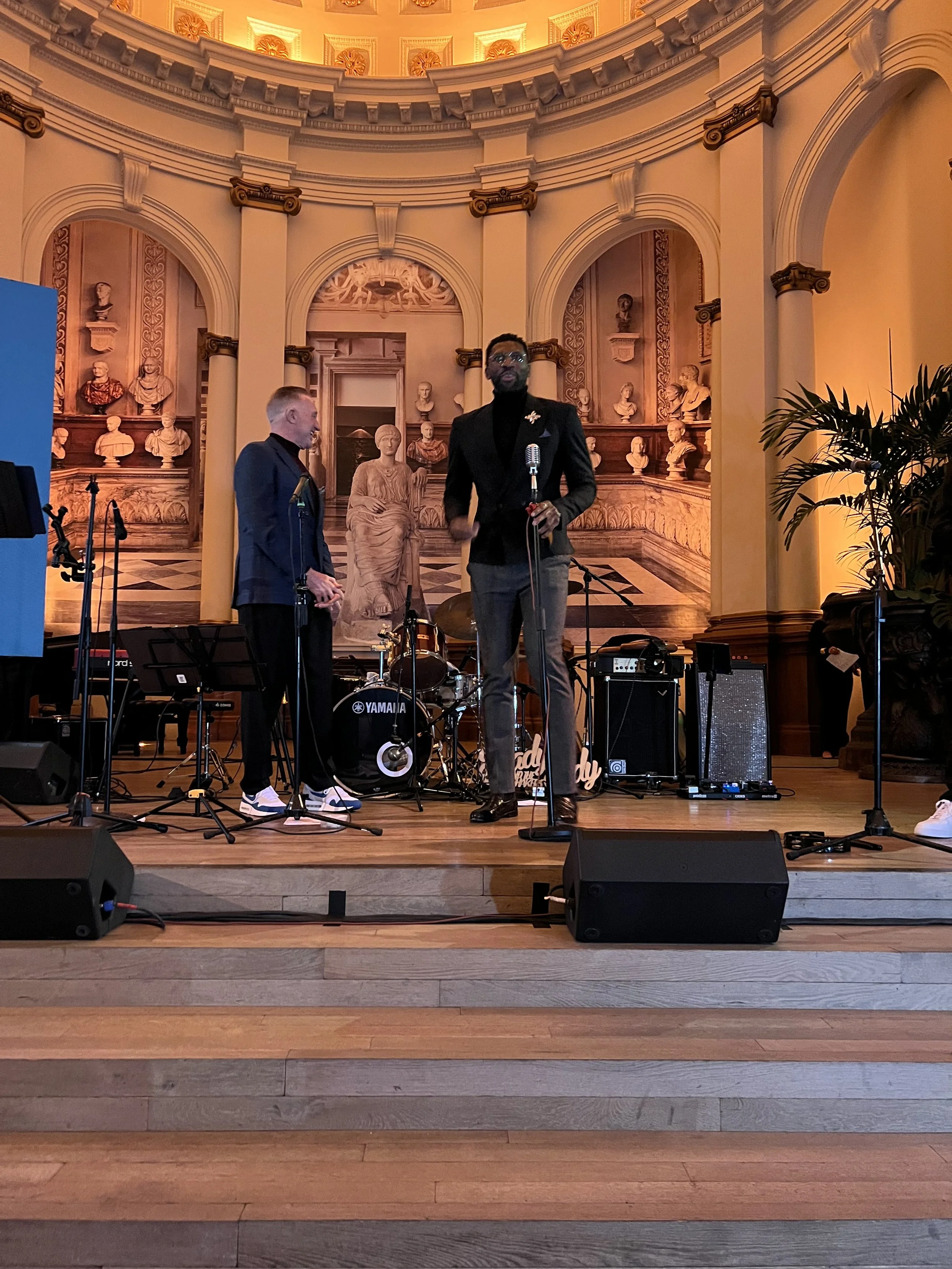 Two men on stage in a grand, ornate hall with classical bust sculptures in the background. One man is speaking into a microphone, wearing a black blazer and gray pants. The other man is standing beside him, wearing a dark blazer and black pants. Musi