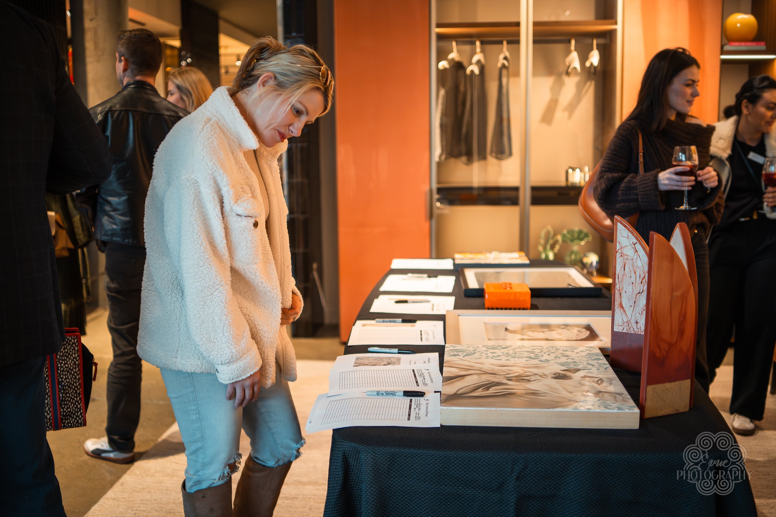 A woman in a cream-colored, fluffy jacket is examining artwork and documents displayed on a black table at an art exhibit. Other people are in the background, some holding glasses of wine.