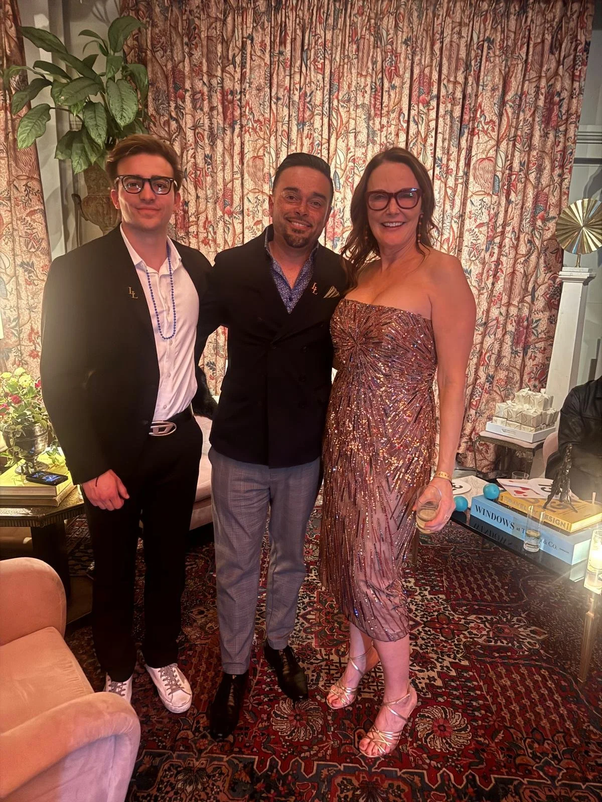 Three people standing together indoors at a formal event, dressed in elegant attire, with patterned curtains in the background and a floral rug on the floor.