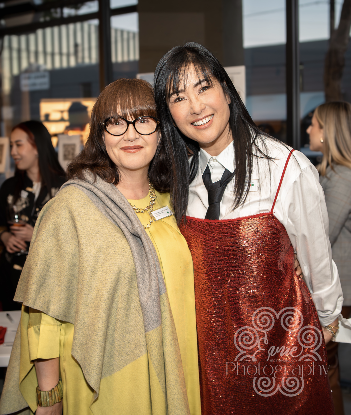 Two women, one with glasses and the other with long black hair, smiling at an indoor event. The woman on the left wears a yellow dress, a cape, and a necklace, while the woman on the right is dressed in a white shirt with a black tie and a red sequin