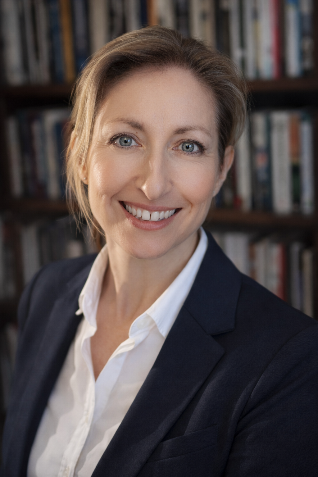 A woman with short blonde hair, wearing a dark blazer and white shirt, smiling at the camera against a blurred bookshelf background in an indoor setting.