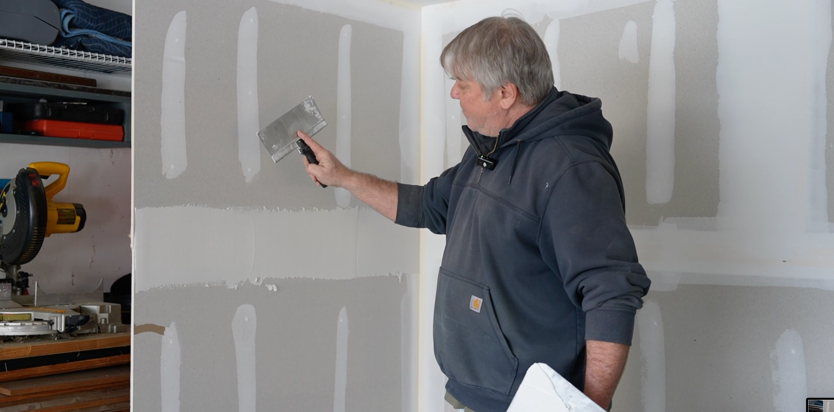 Man in a dark hoodie measuring and patching drywall in a room.