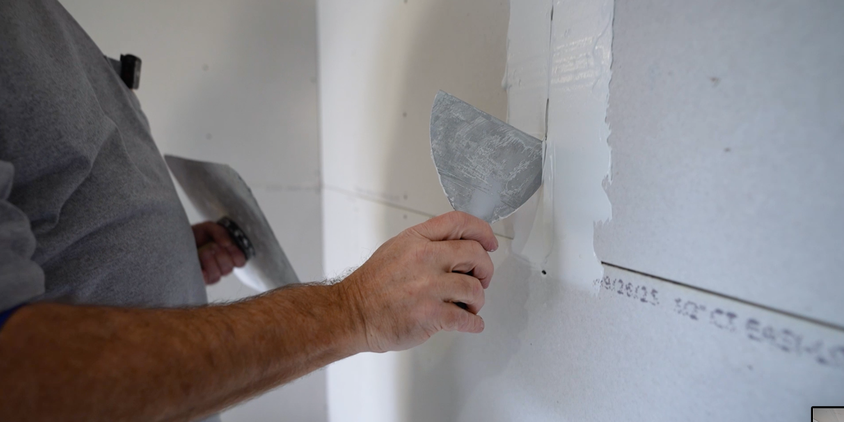 A person's hand is applying joint compound to a wall using a putty knife during drywall installation.