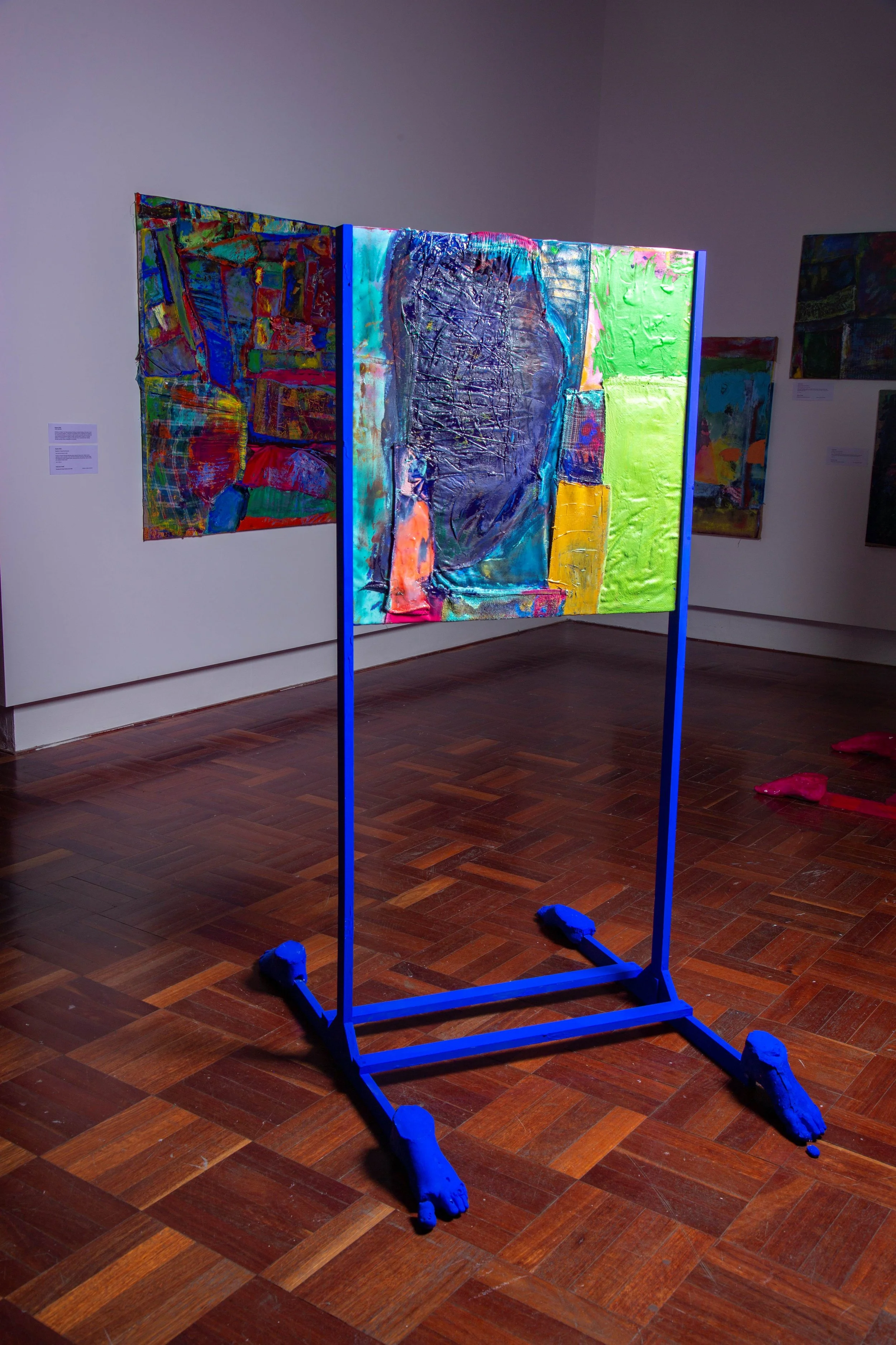 Colorful abstract painting on an easel in an art gallery, with other artworks on the walls, and a polished wooden floor.