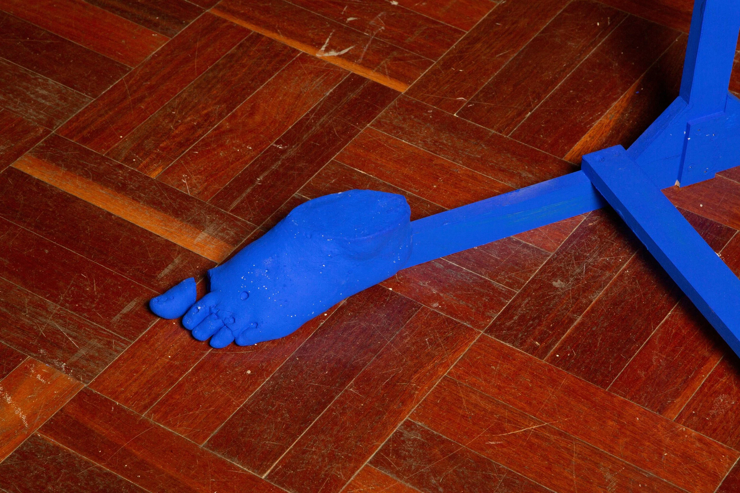 A blue-painted wooden sculpture of a foot, lying on a reddish-brown wooden floor.