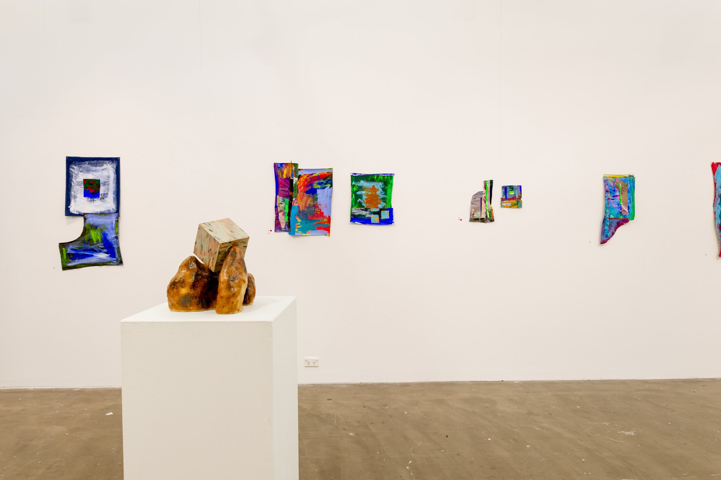 Contemporary art gallery with colorful abstract paintings on a white wall and a sculpture of a hand holding a cube on a pedestal.