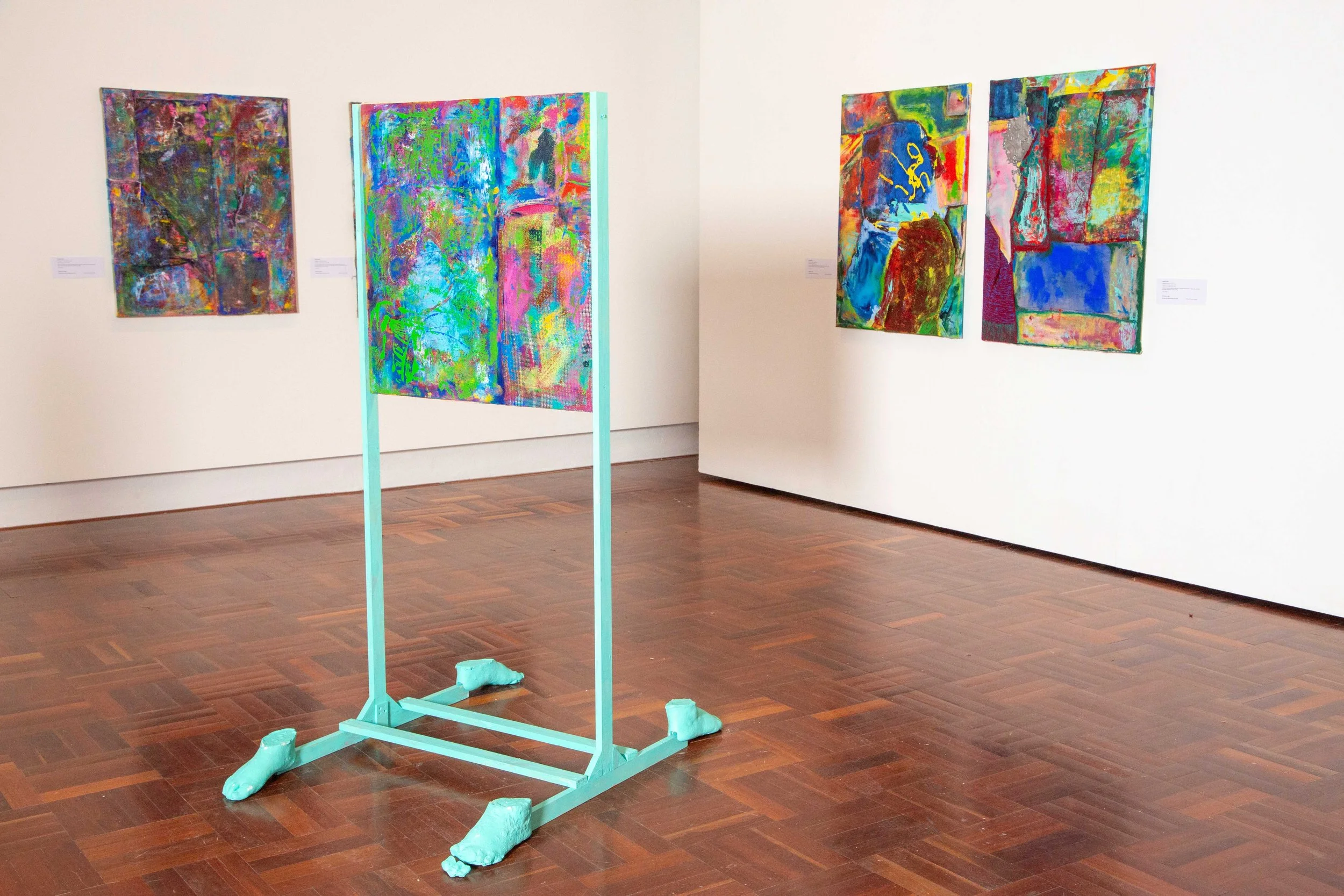 An art gallery featuring four abstract paintings on white walls and a turquoise frame on the wooden floor. The frame holds fabric resembling two shoes.
