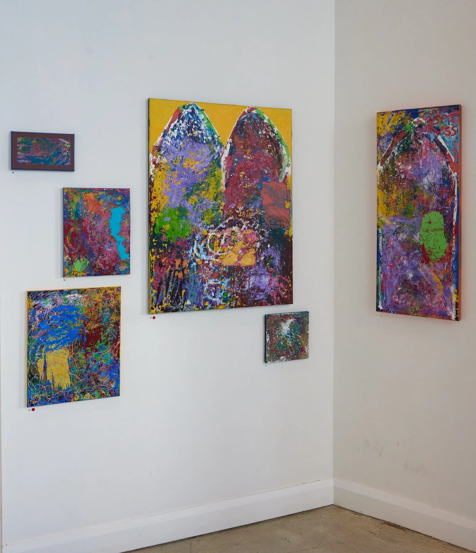 Gallery wall with abstract colorful paintings, including a large piece with two mountain-like shapes and smaller paintings with splashes of bright colors.