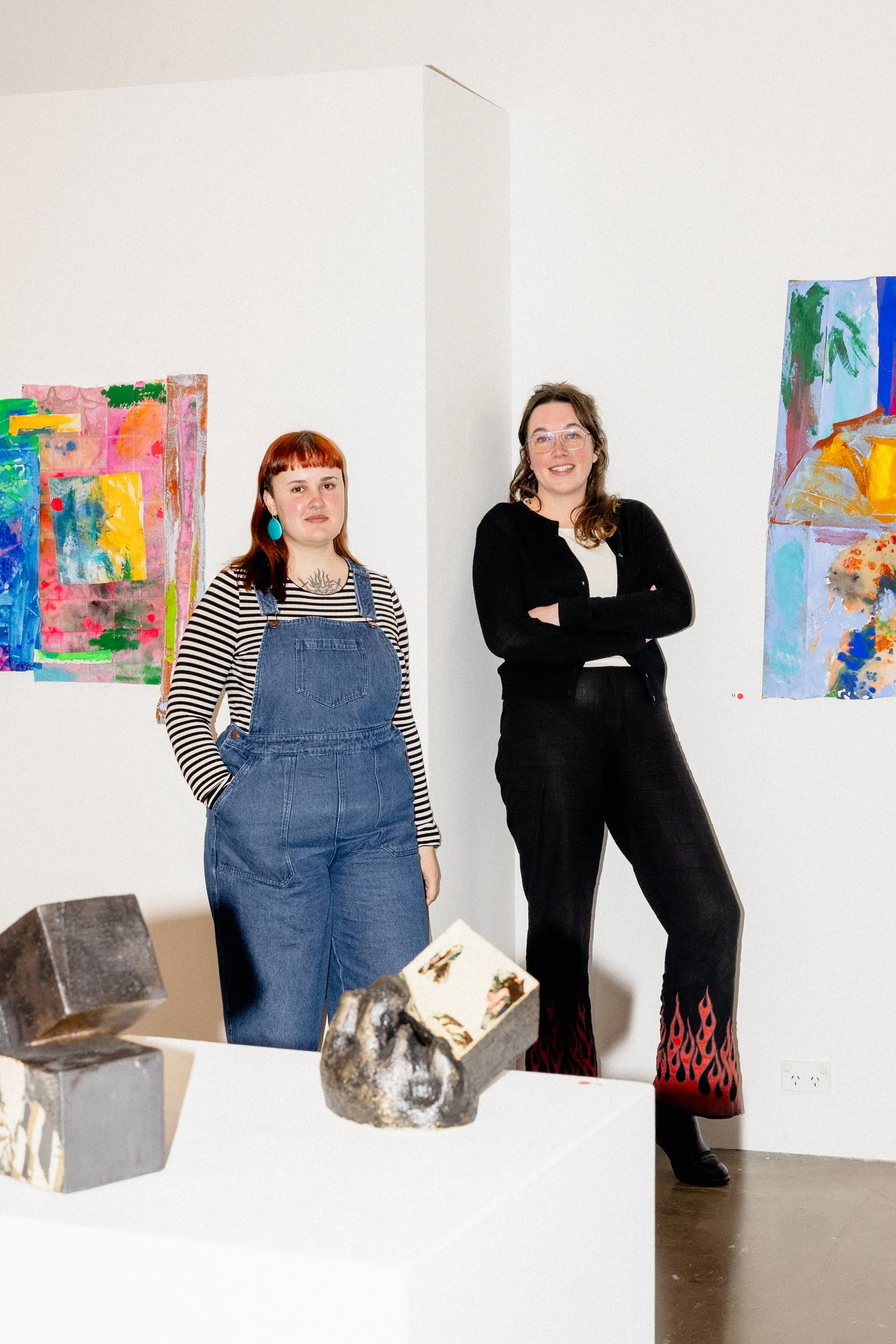 Two women stand in an art gallery with colorful abstract paintings on the white wall behind them and sculptures on a white pedestal in the foreground.