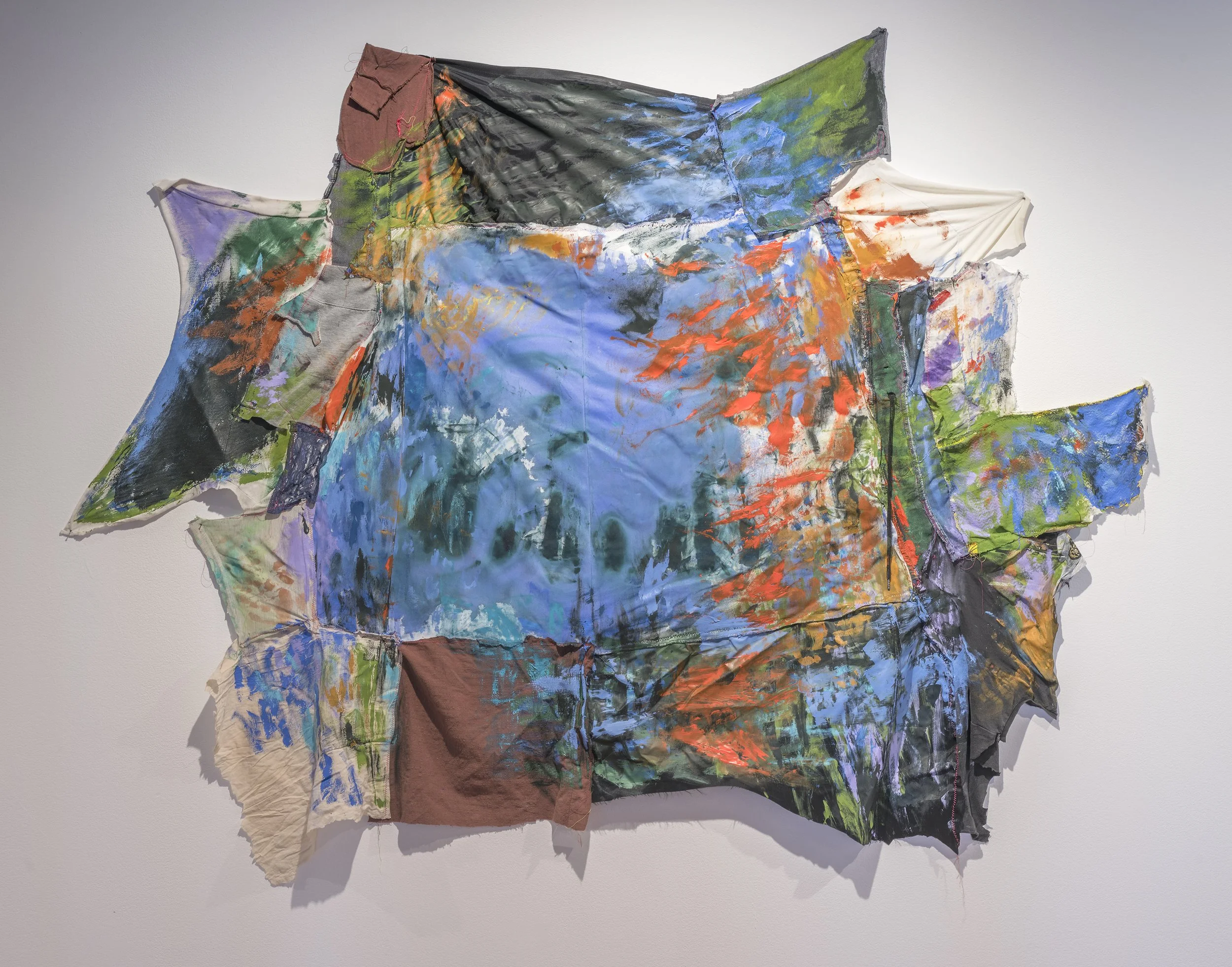 Mixed media artwork consisting of painted, fabric, and textile pieces arranged on a wall, featuring abstract shapes and vibrant colors.