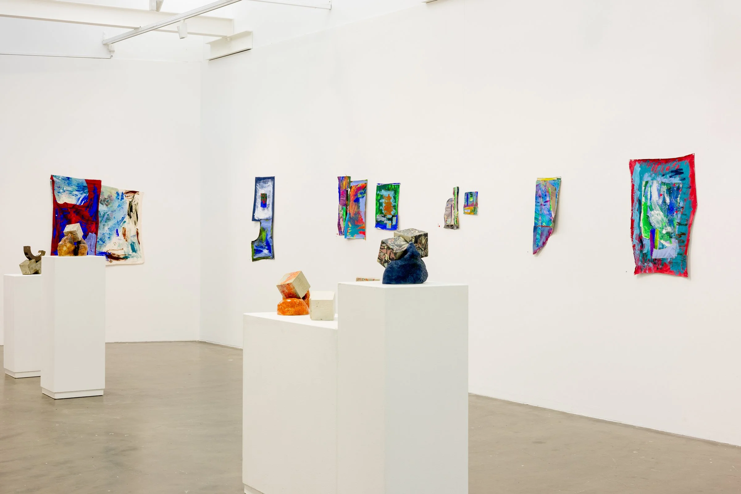 An art gallery with white walls and concrete floors showcasing colorful abstract artworks on the walls and ceramic sculptures on white pedestals.