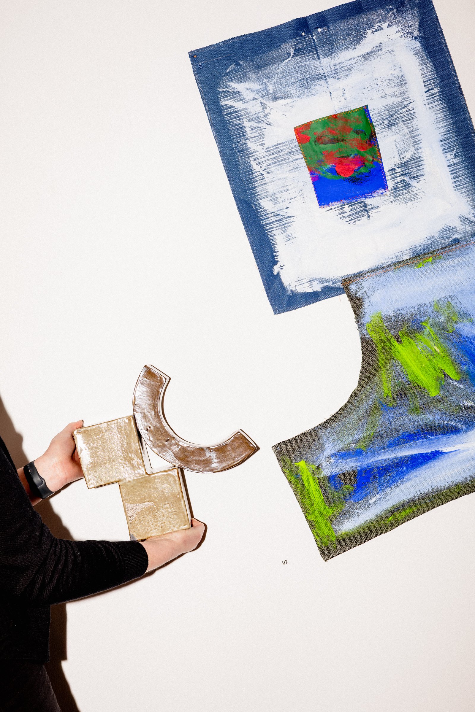 A person holding a metallic T-shaped object and a semi-circular object in front of an abstract painting with blue, green, white, red, and brown colors on a white wall.